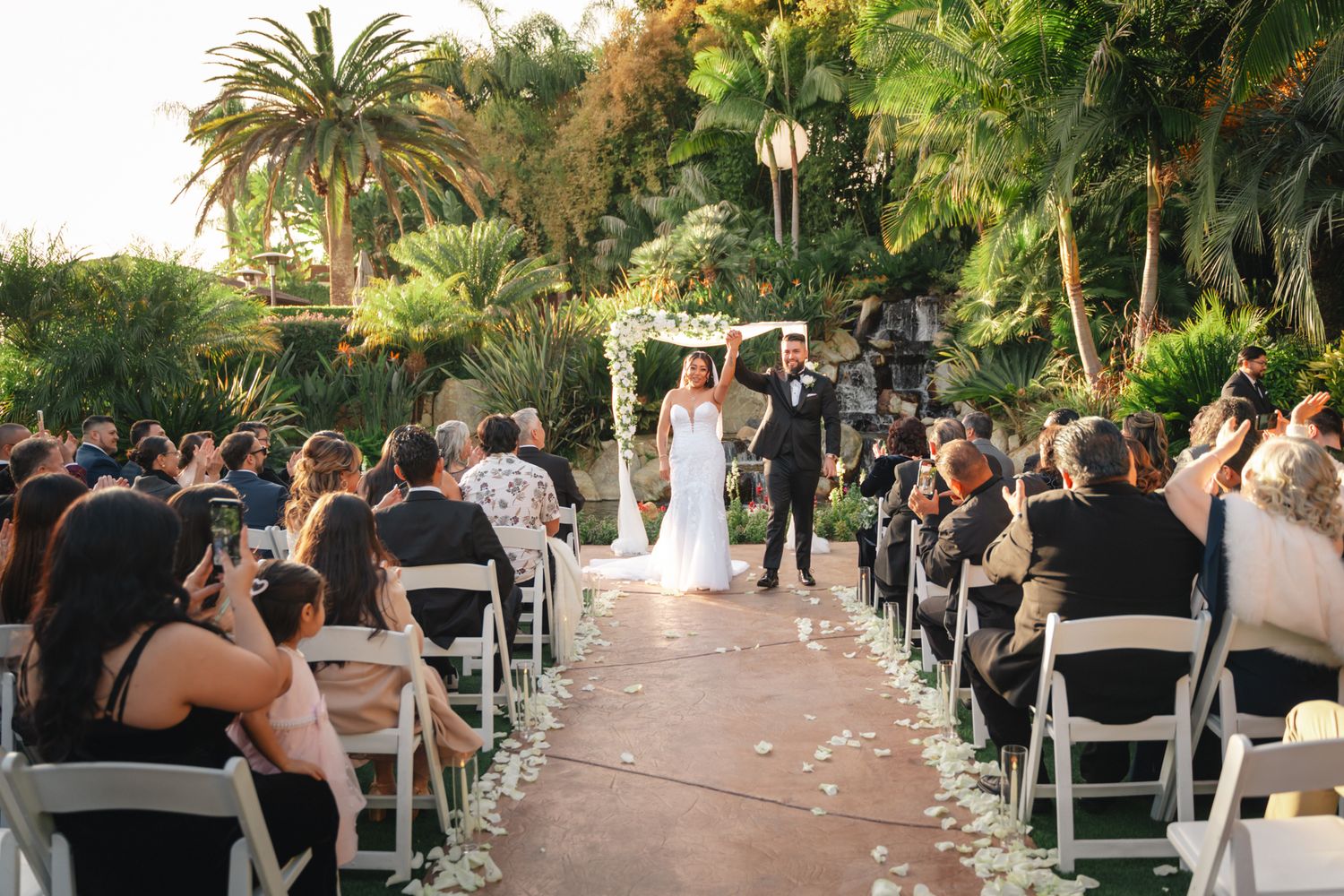 Wedding ceremony guests seated in white chairs along a palm tree-lined pathway at an outdoor venue during sunset.