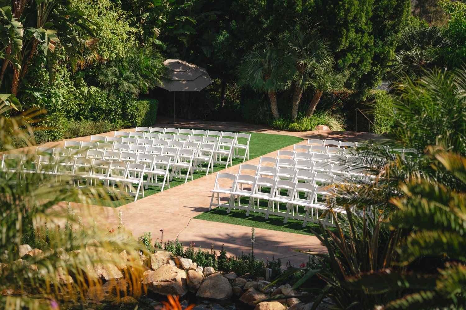 White ceremony chairs arranged in rows along a garden path surrounded by lush greenery and natural landscaping.