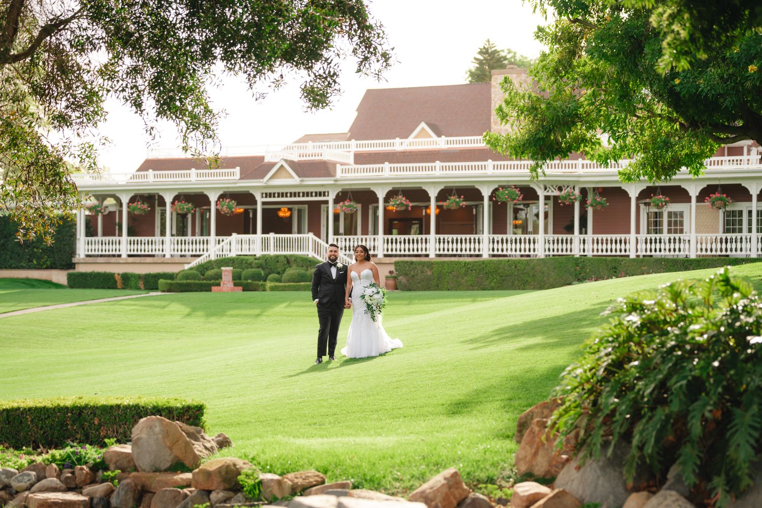 Elegant wedding couple on manicured lawn in front of historic Victorian mansion with ornate white wraparound porch.