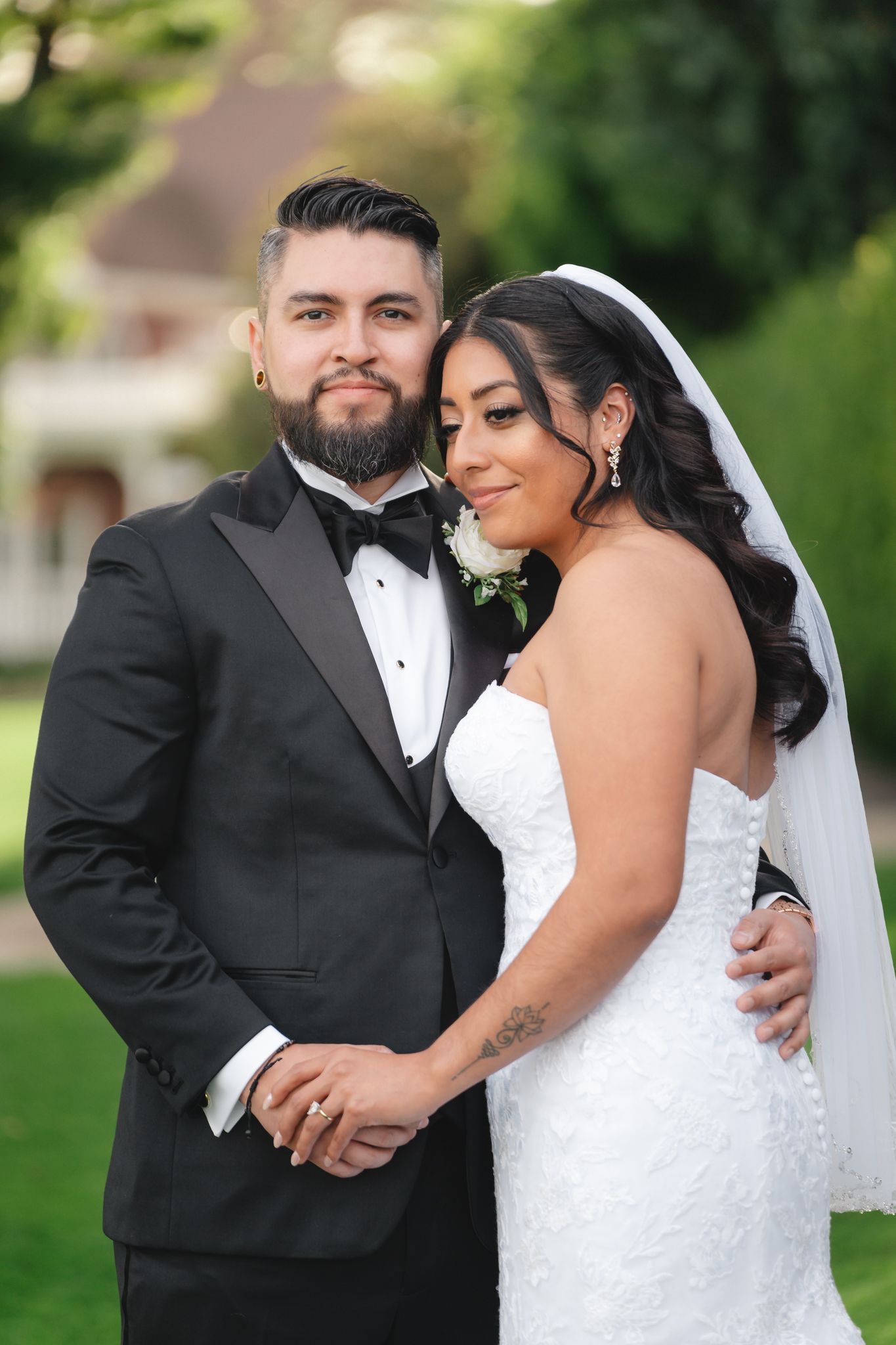 A romantic wedding couple embraces outdoors in formal attire against a green garden backdrop.