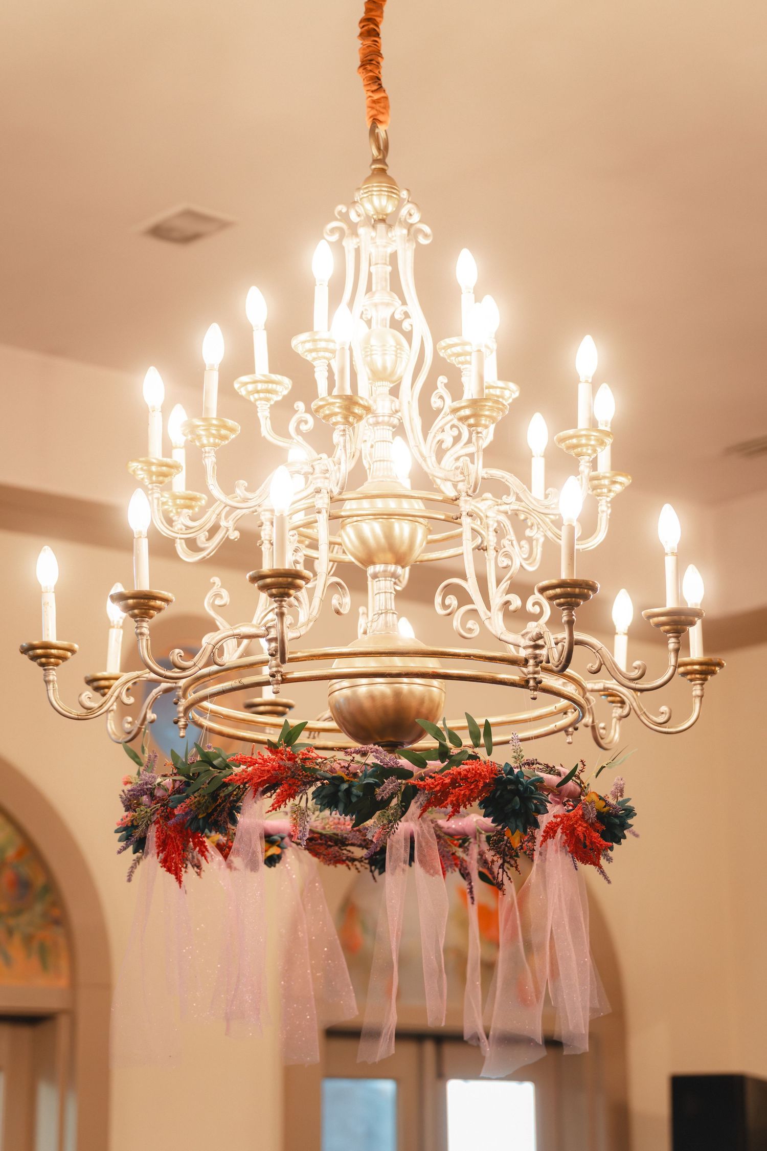 Elegant gold chandelier with candle lights decorated with colorful flowers and pink ribbons hanging from ceiling.