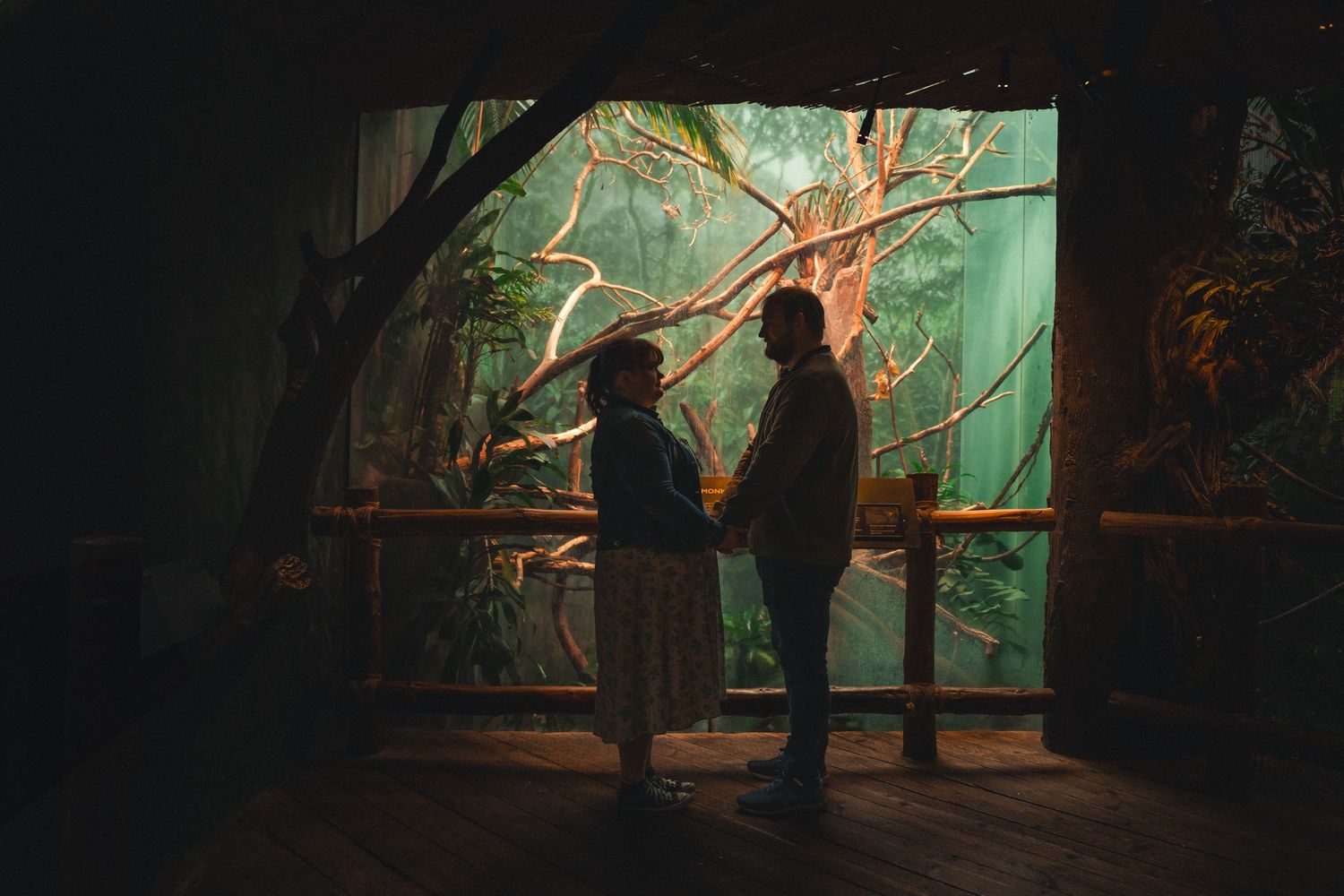 Two silhouettes stand at a railing in front of an illuminated aquarium display with glowing turquoise water.