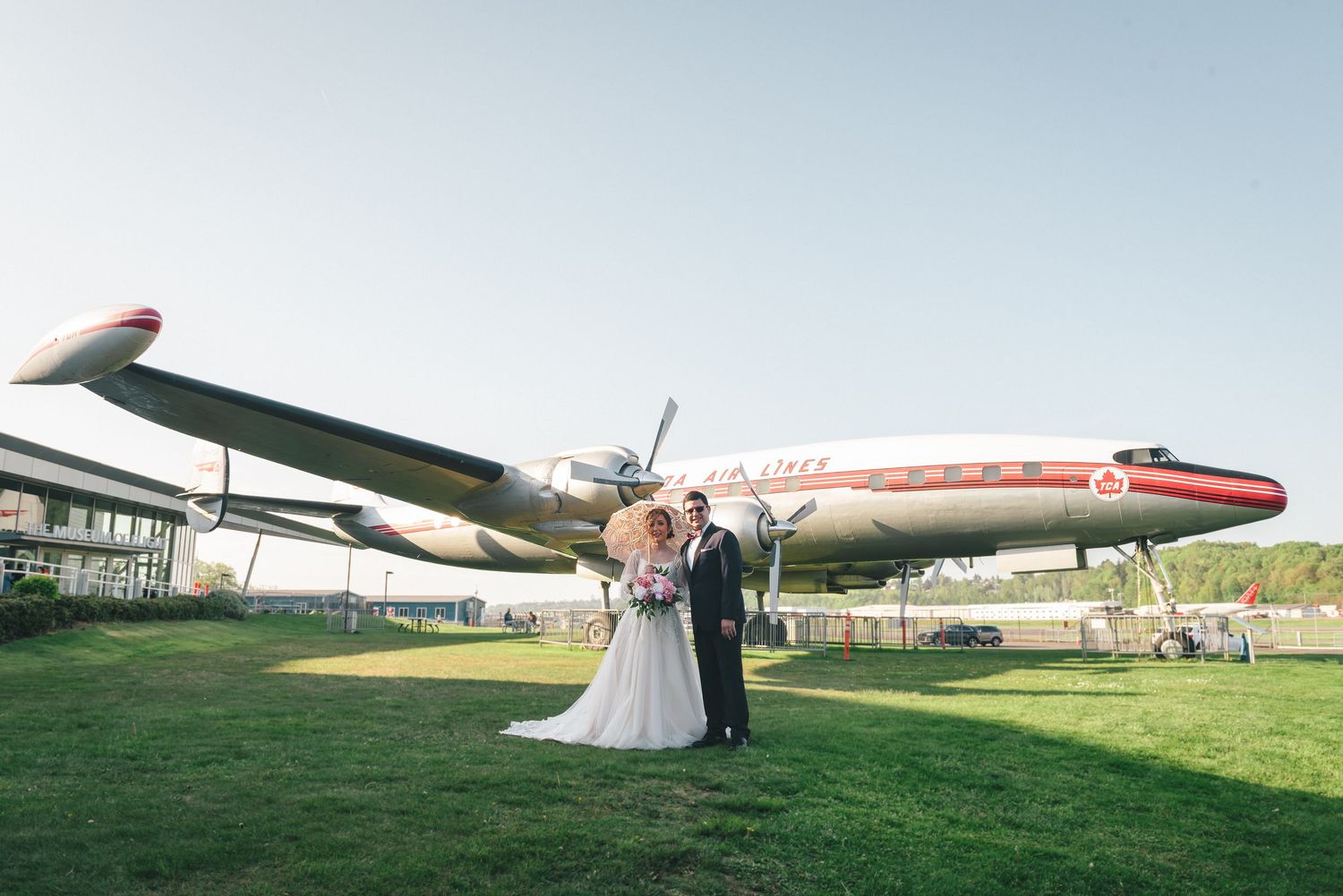 A newlywed couple poses in front of vintage aircraft at an outdoor aviation museum during their wedding photoshoot.