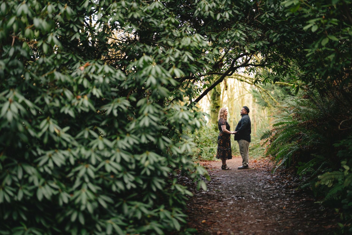 Two figures walk down a shaded forest path surrounded by lush green foliage and trees.