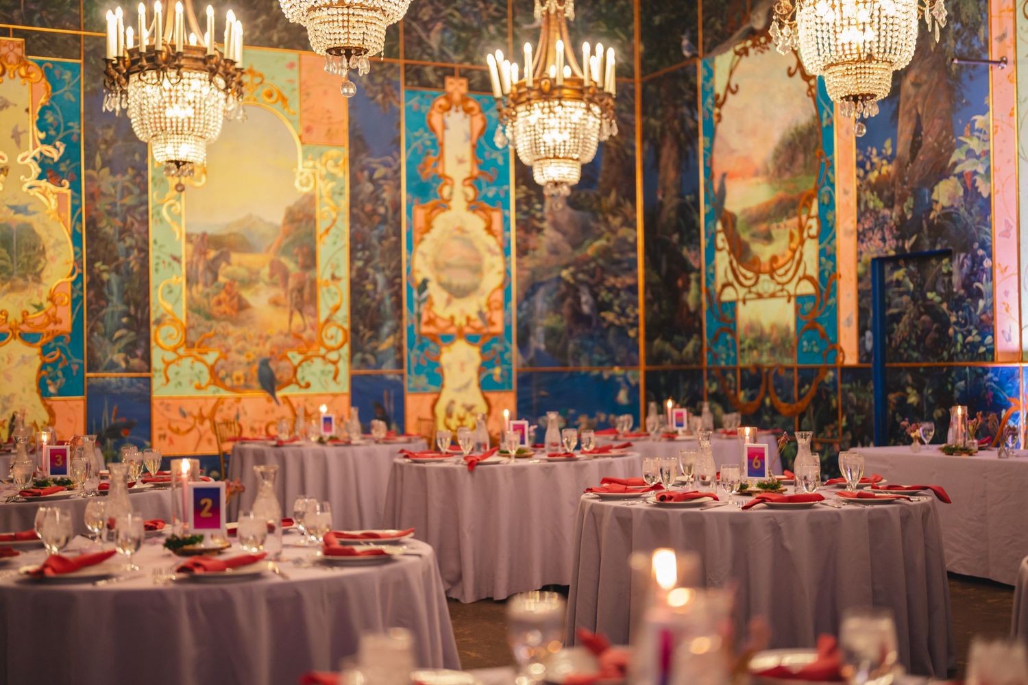 Elegant banquet hall with crystal chandeliers, ornate murals, and round tables set with red napkins and glassware.