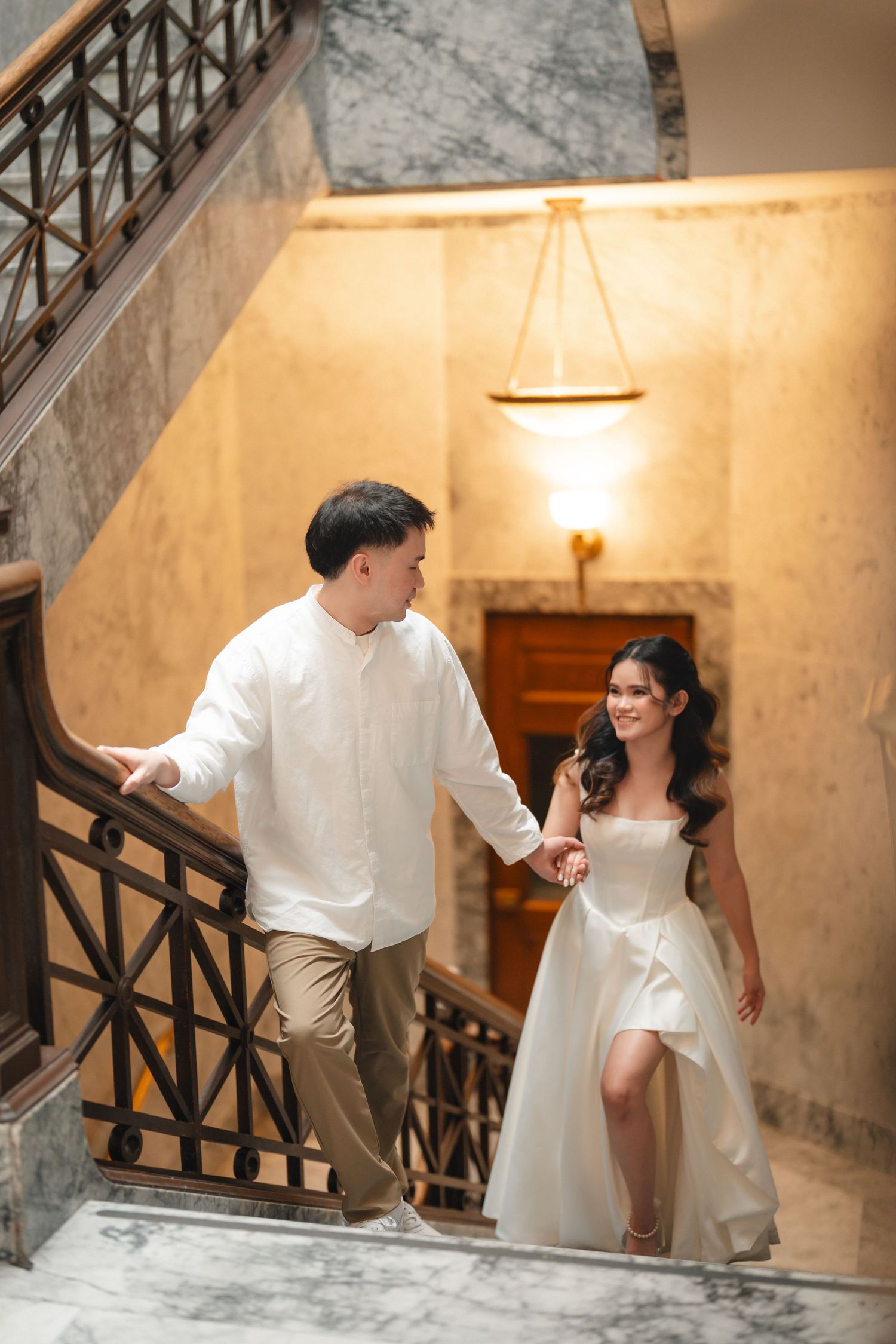 A couple in white formal attire walking up elegant marble stairs with ornate railings and warm lighting.