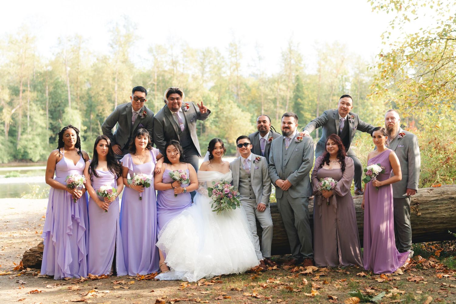 Wedding party in purple and gray attire poses outdoors near a lake during autumn.