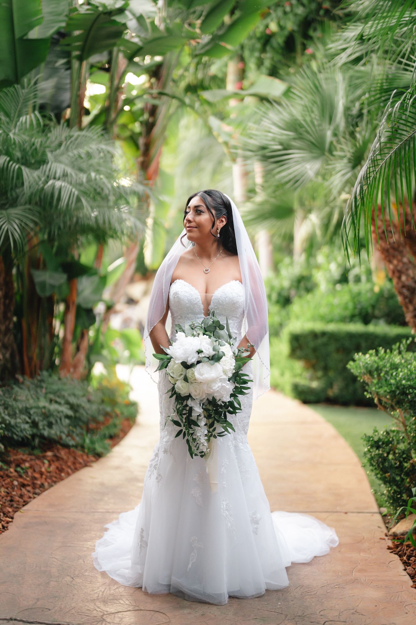 A white wedding dress with lace details photographed in a lush tropical garden setting with palm trees and a winding path.