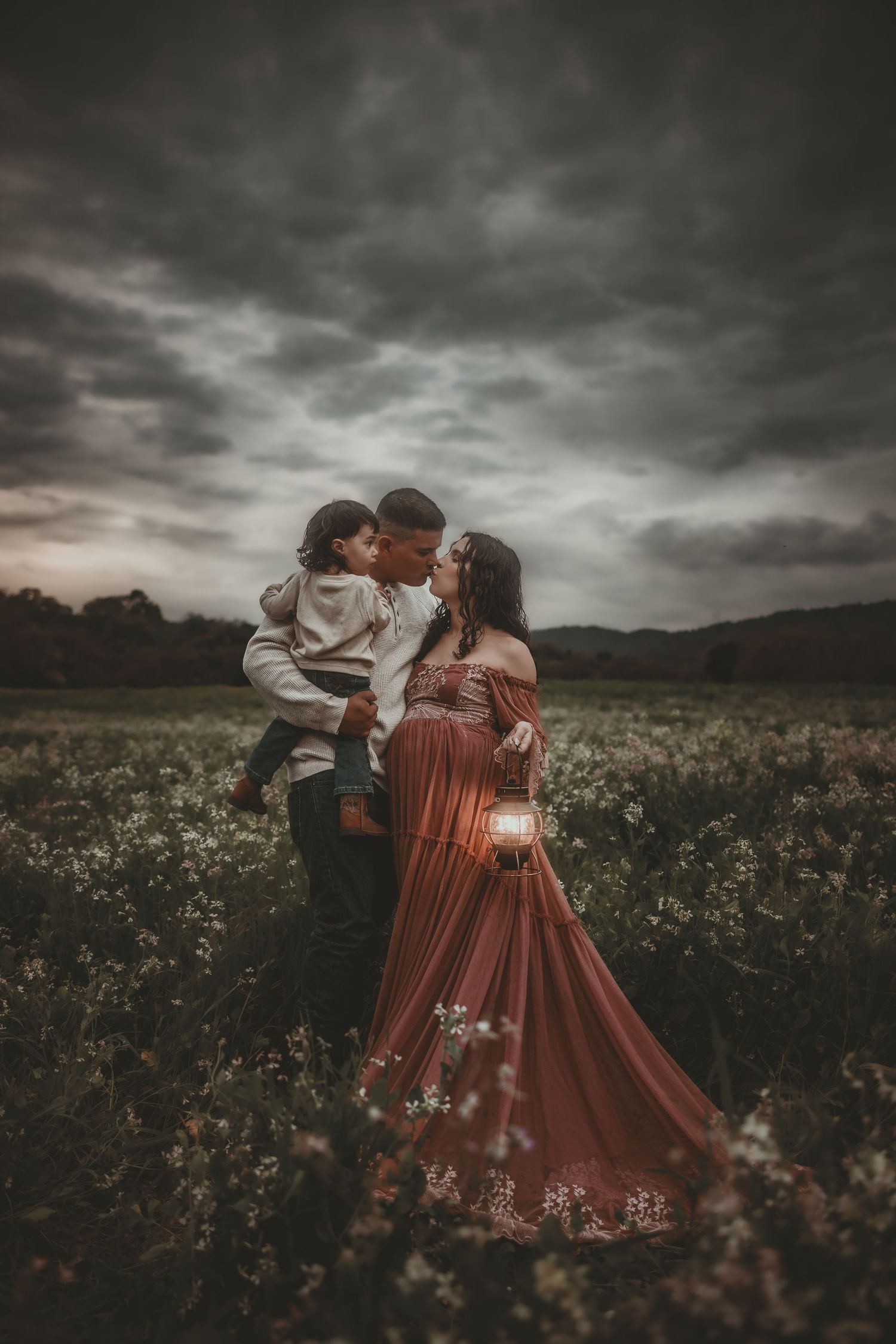 A romantic expecting couple embraces with their child in a wildflower field under dramatic stormy skies wearing vintage-inspired clothing.
