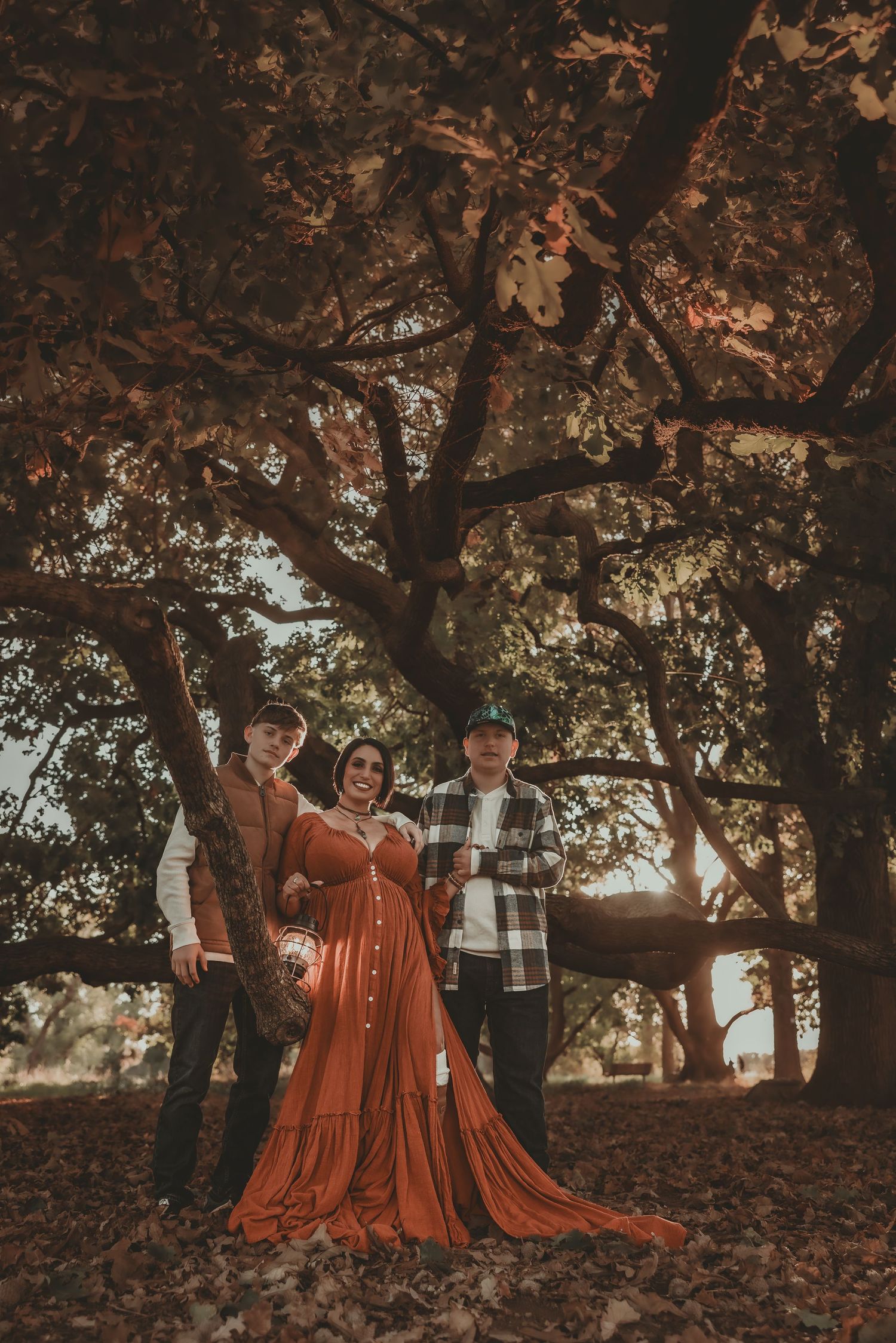 Family maternity photo under autumn oak tree, woman in flowing rust dress with two males in fall foliage setting.