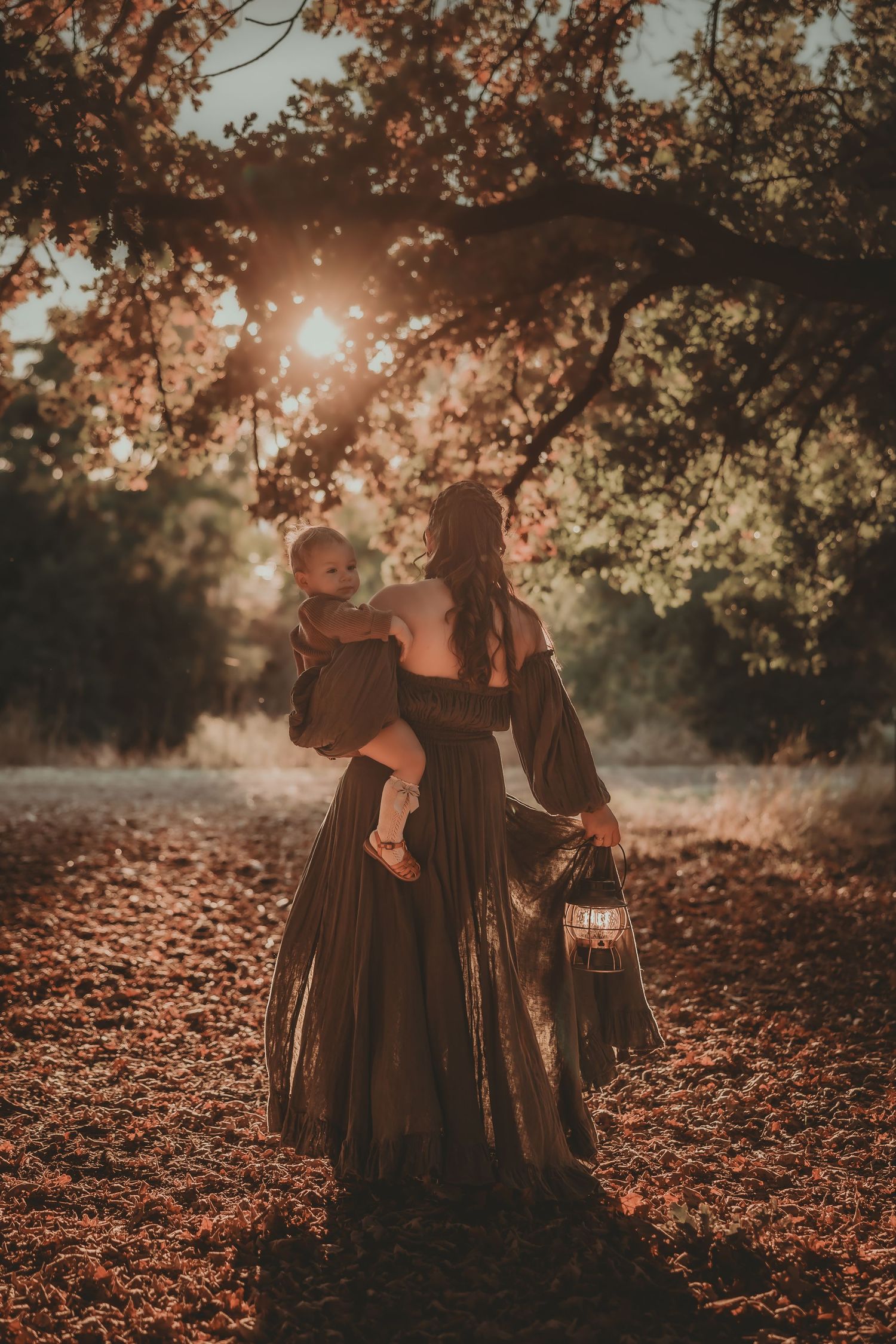 Woman in dark gown holds baby under sunlit autumn trees, carrying a lantern in a dreamy forest setting.