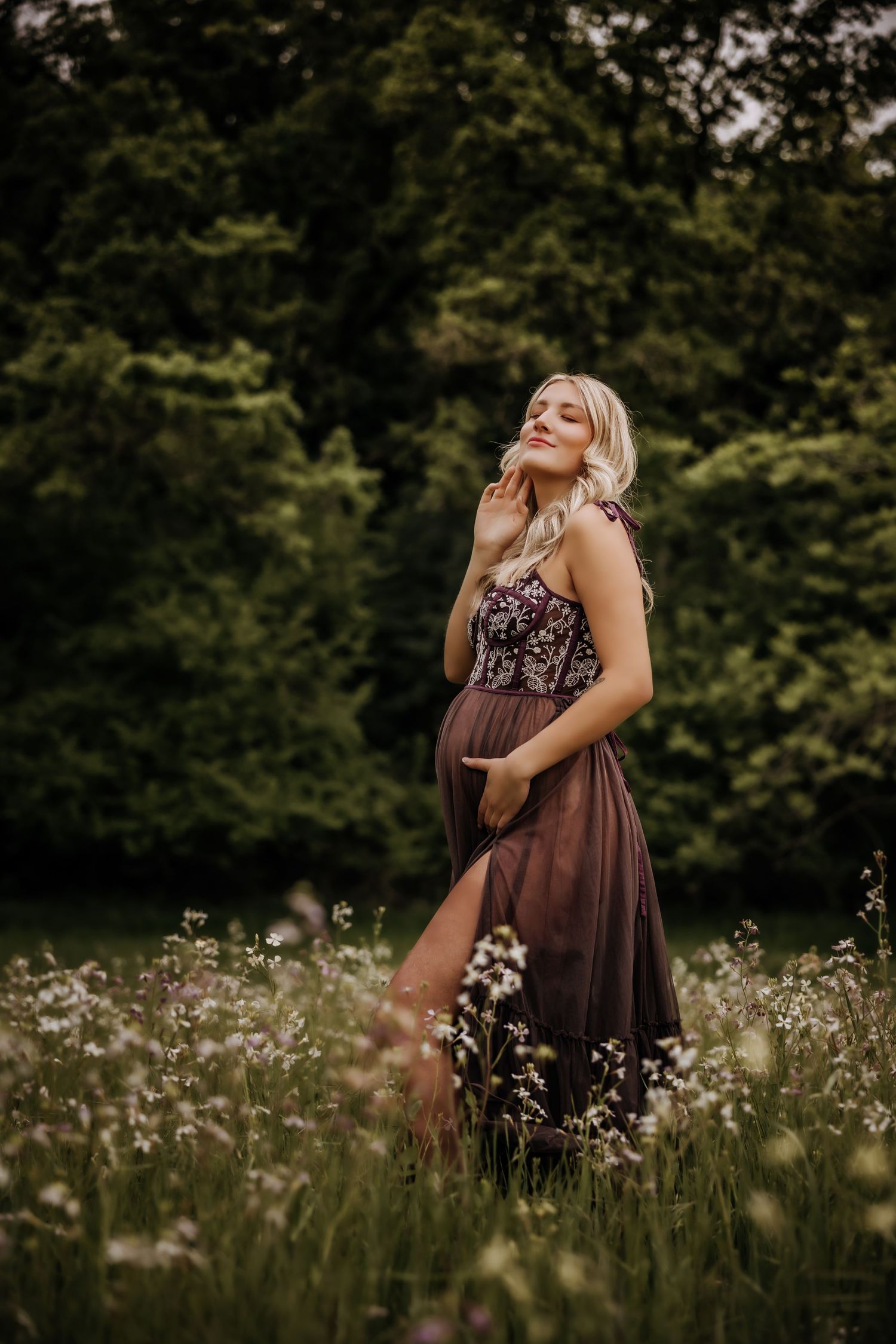 A woman in a floral dress stands in a wildflower meadow at sunset with trees in the background.