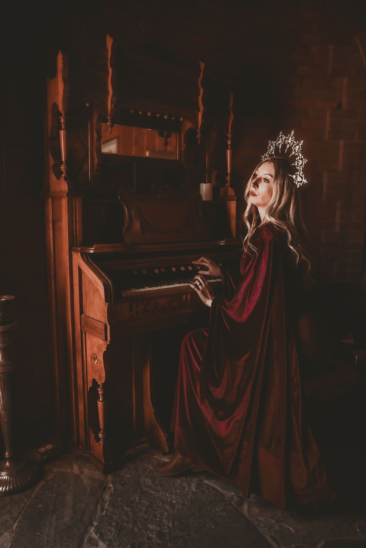 A woman in a red velvet gown and silver crown plays an antique wooden piano in a dark, medieval-style room.