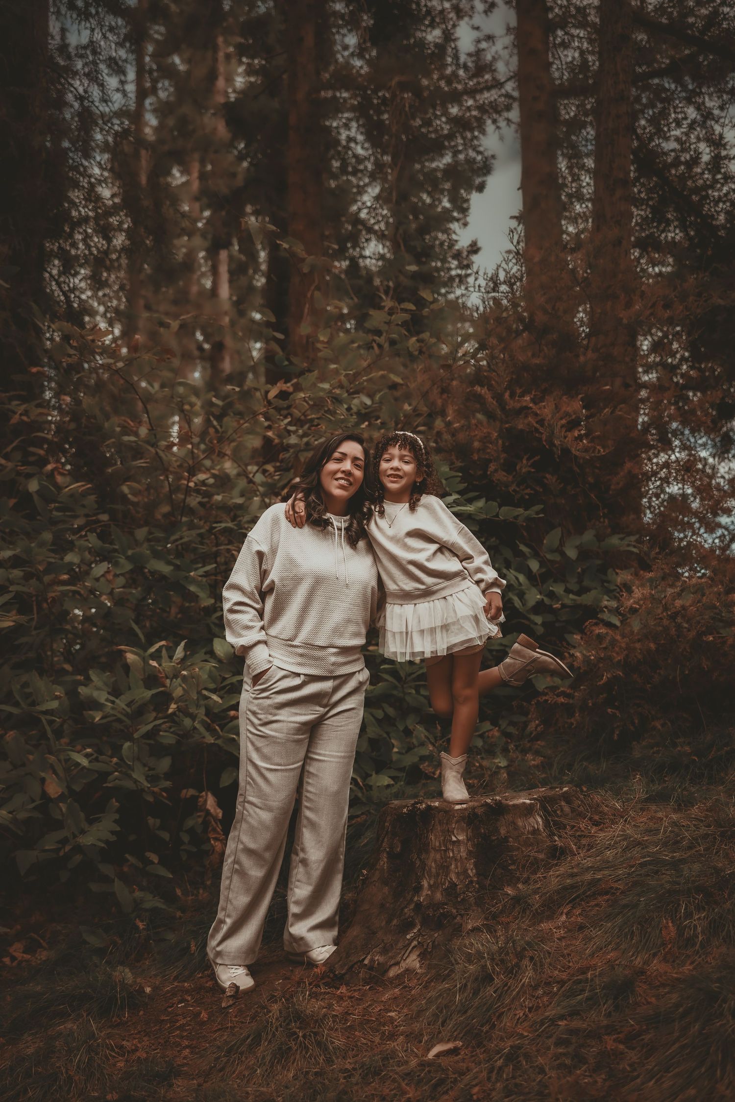 A woman and young girl in matching cream outfits pose together in a lush forest setting.