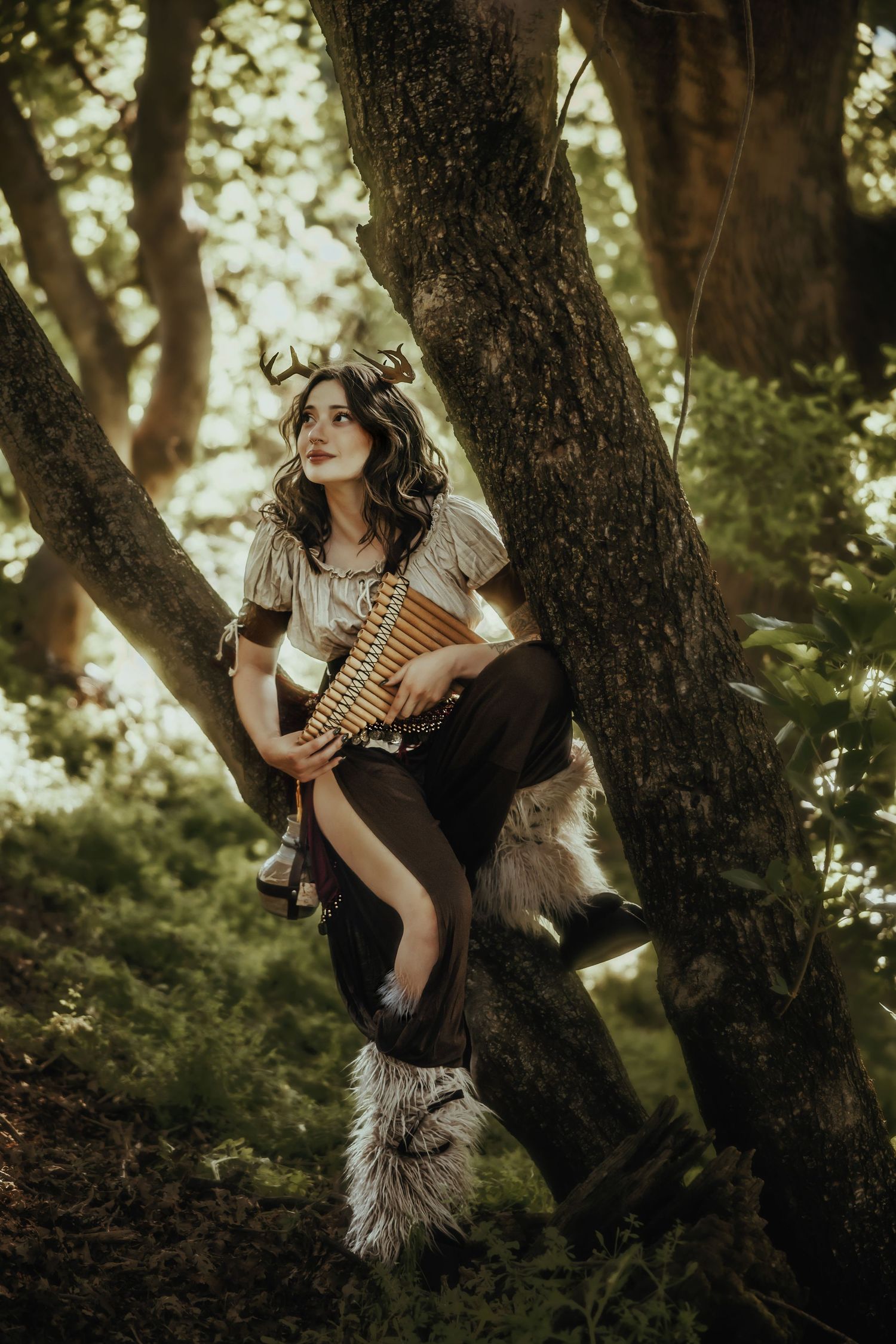 A figure in dark clothing sits perched in a tree branch while playing a pan flute in a mystical forest setting.