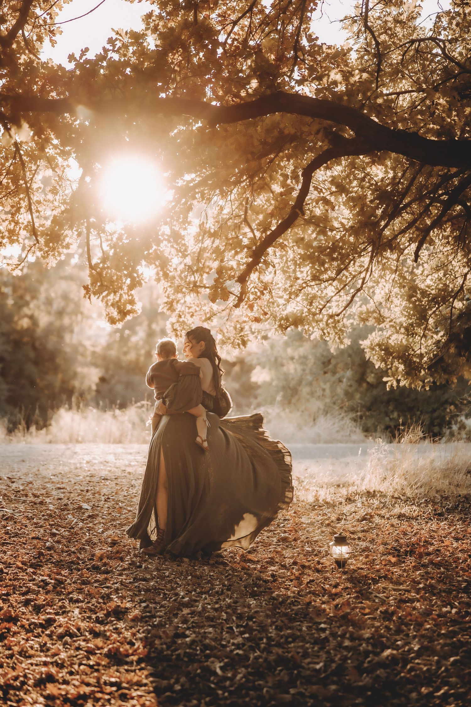 A mother in a flowing olive dress holds her baby under a golden oak tree at sunset in an autumn forest.