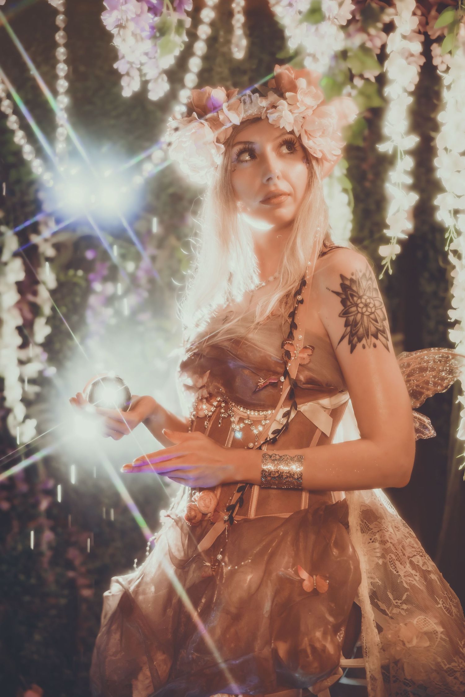 Blonde woman in fairy costume with floral crown holds glowing orb amid sparkling lights and hanging flowers.