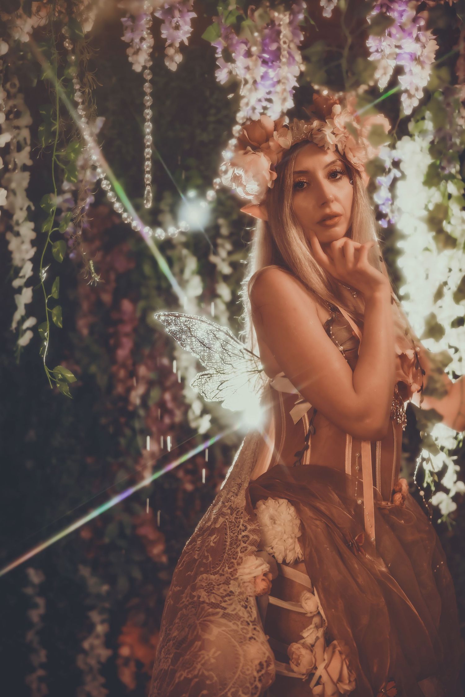 Blonde woman in fairy costume with floral crown, lace dress, and wings posing among hanging flowers and lights.