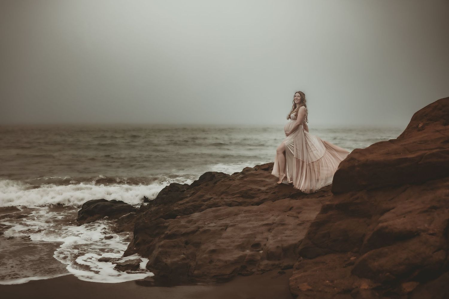A person in a flowing pink dress poses gracefully on dramatic coastal cliffs as ocean waves crash below.