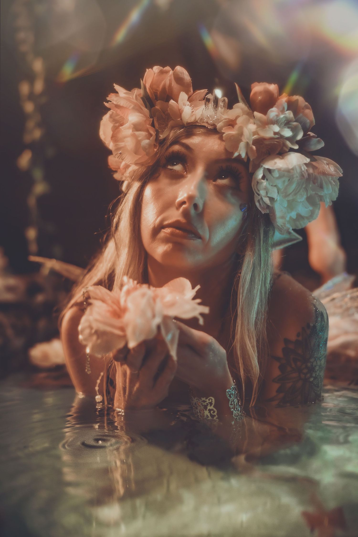 Woman with floral crown in water holding pink flowers, soft bokeh light effects, dreamy fantasy portrait photography.