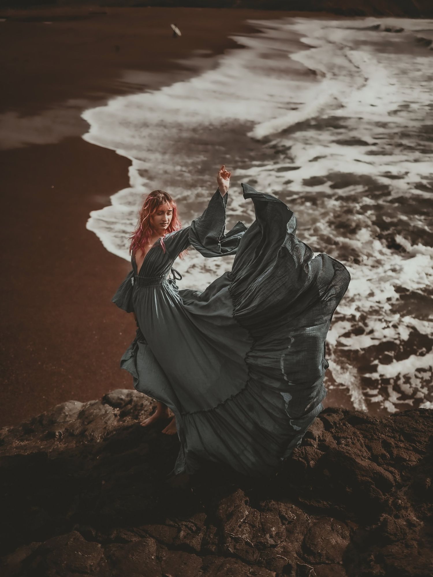 A woman in a flowing dark gown stands on rocky shore, red hair blowing as ocean waves crash behind her.