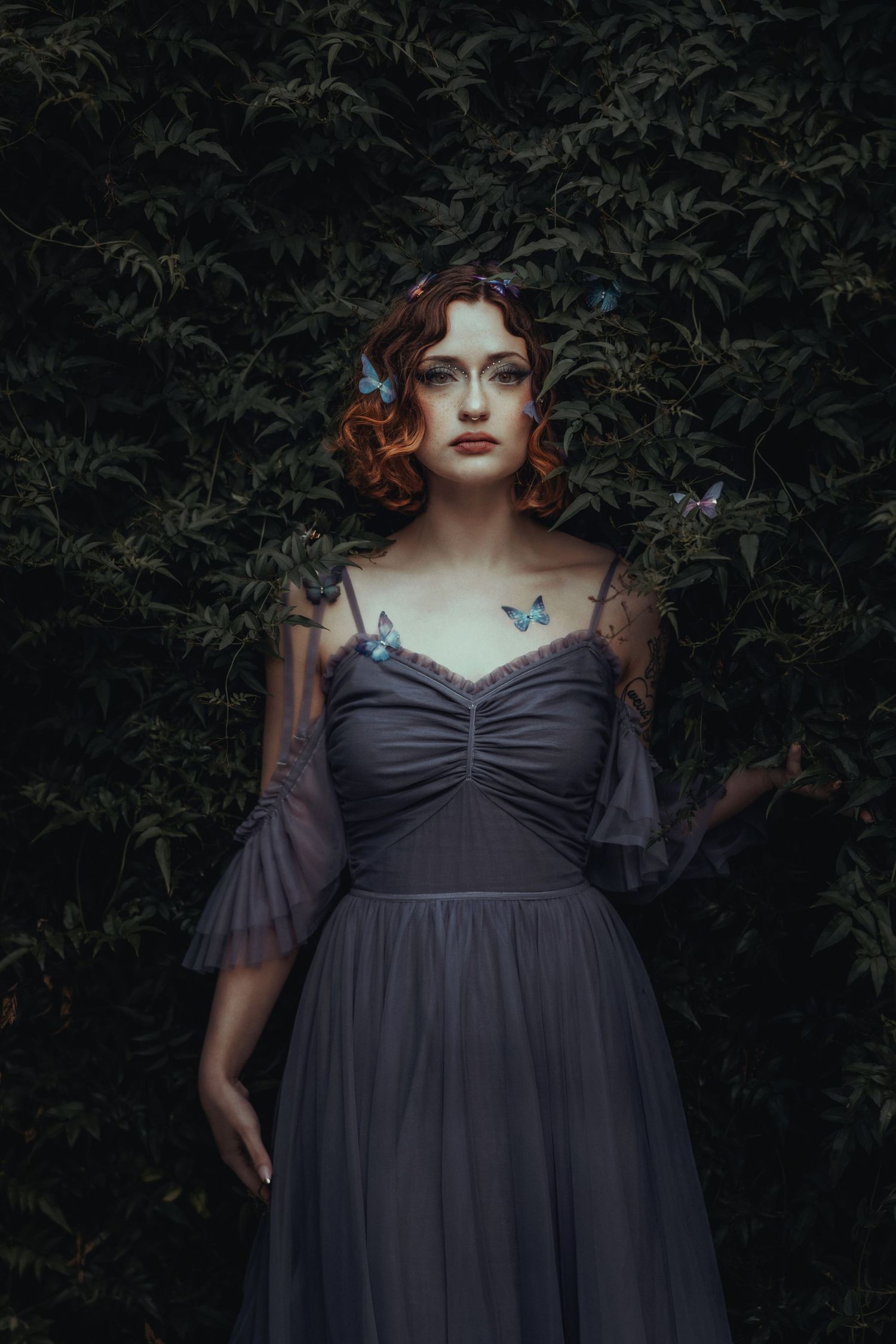 A figure in a dark purple dress poses against a shadowy garden backdrop with delicate blue butterflies.