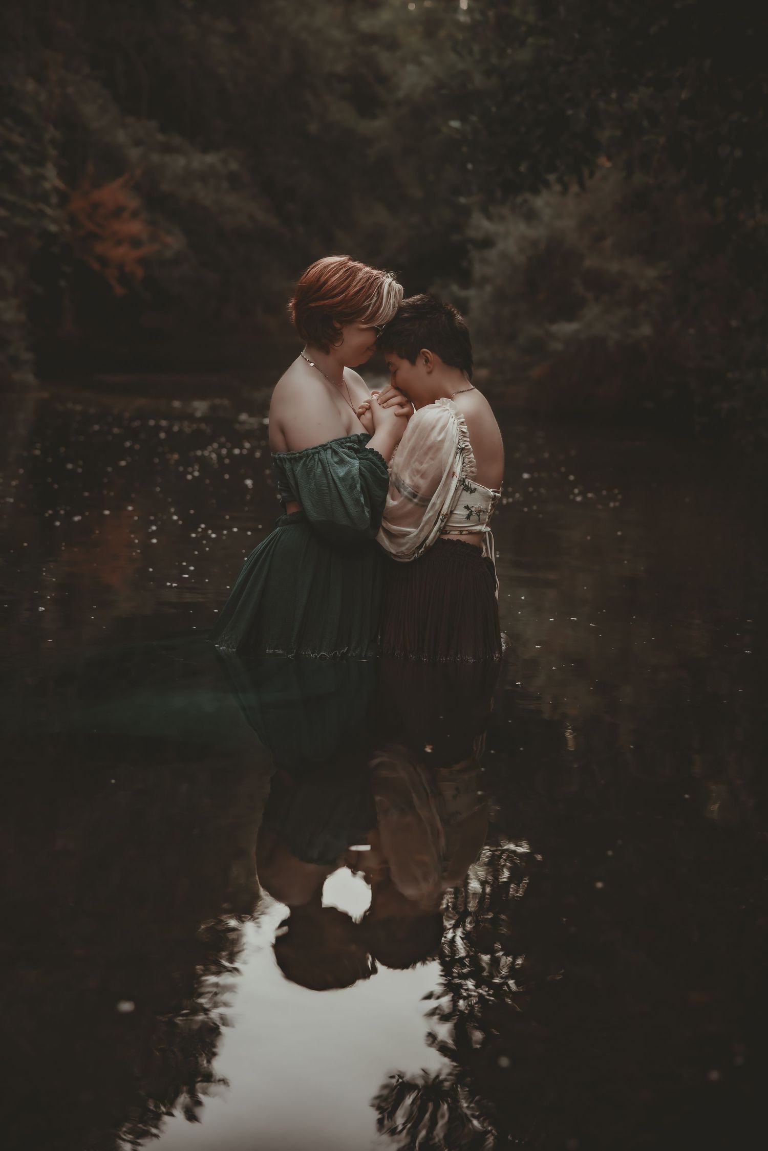 Two women in vintage dresses embrace romantically by a dark reflective forest pond.