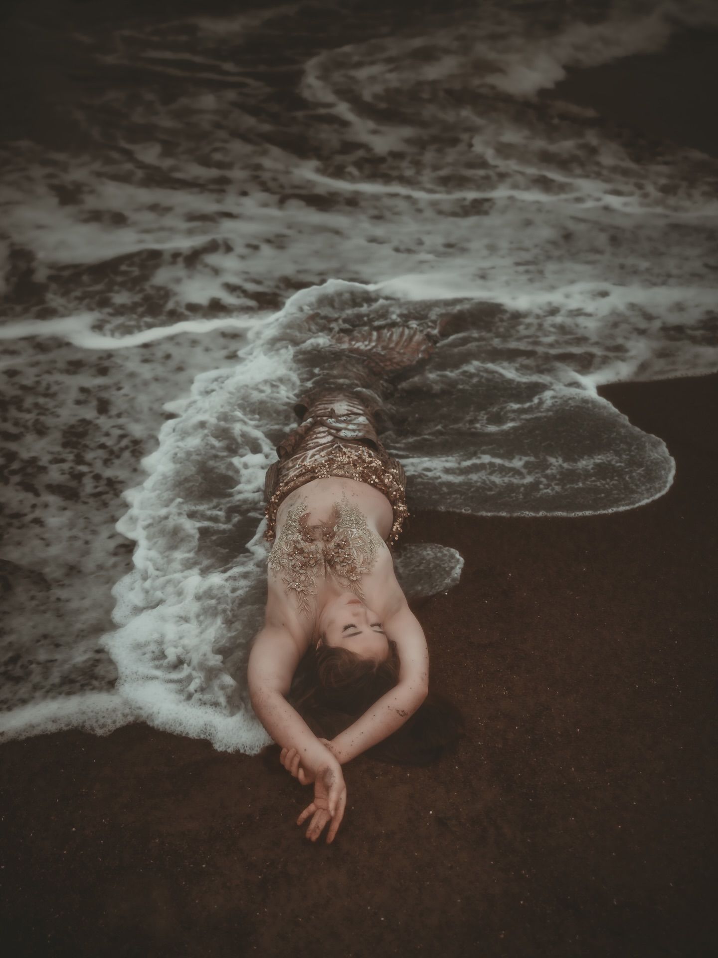 A person lying on a dark sandy beach as ocean waves wash over their body in a dramatic, ethereal scene.