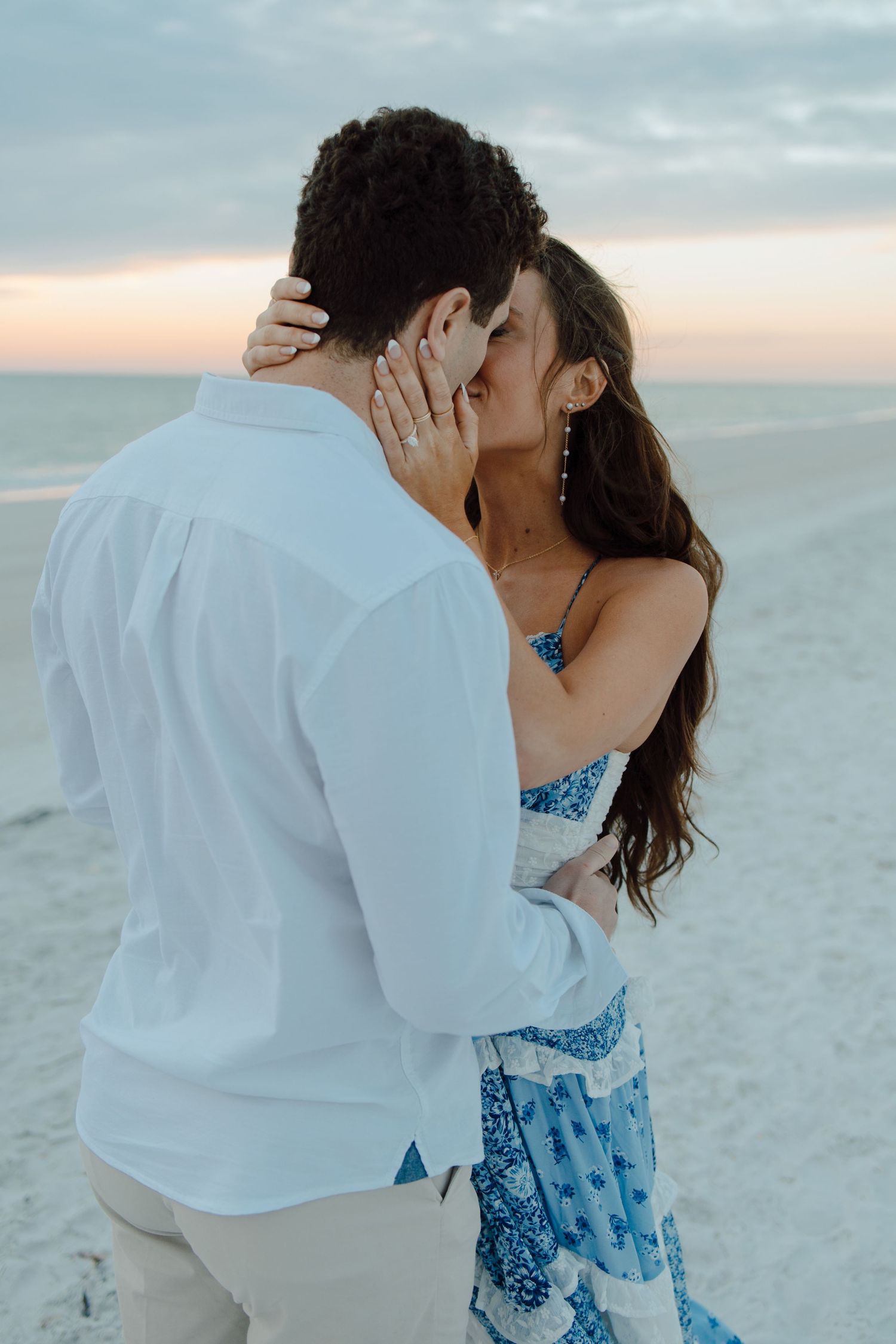 Couple sharing a romantic kiss on a beach at sunset, woman in blue floral dress.