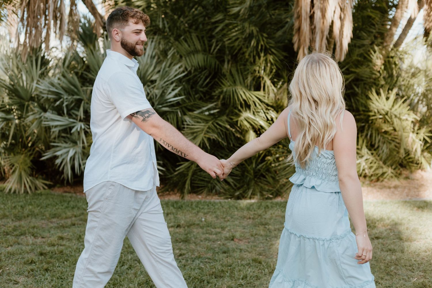 Couple holding hands walking through lush green park, woman in light blue dress, man in white.