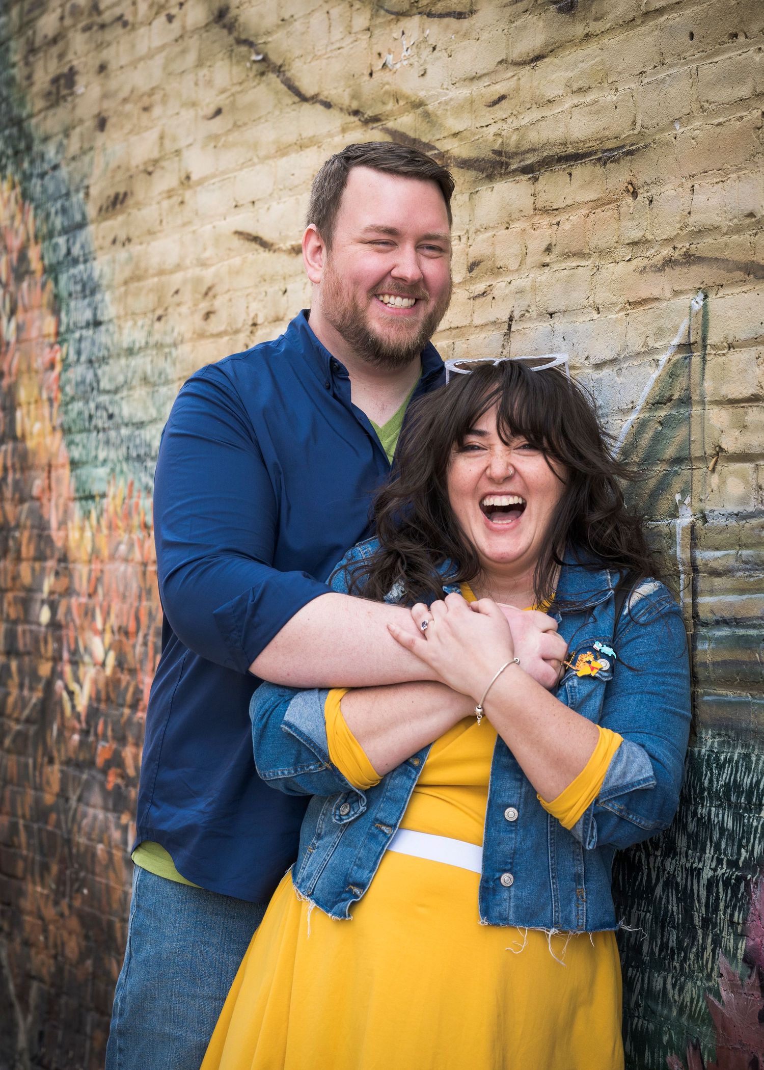 A joyful couple embraces and laughs together against a graffiti wall, with her wearing a yellow dress and denim jacket.