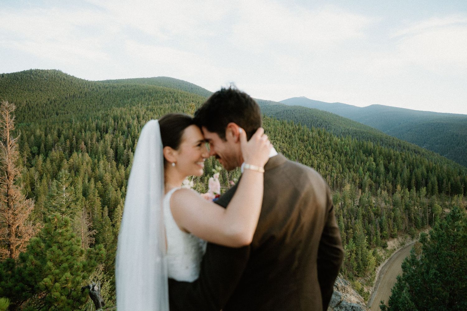 A couple embraces during their wedding photoshoot with evergreen forest mountains in the background.