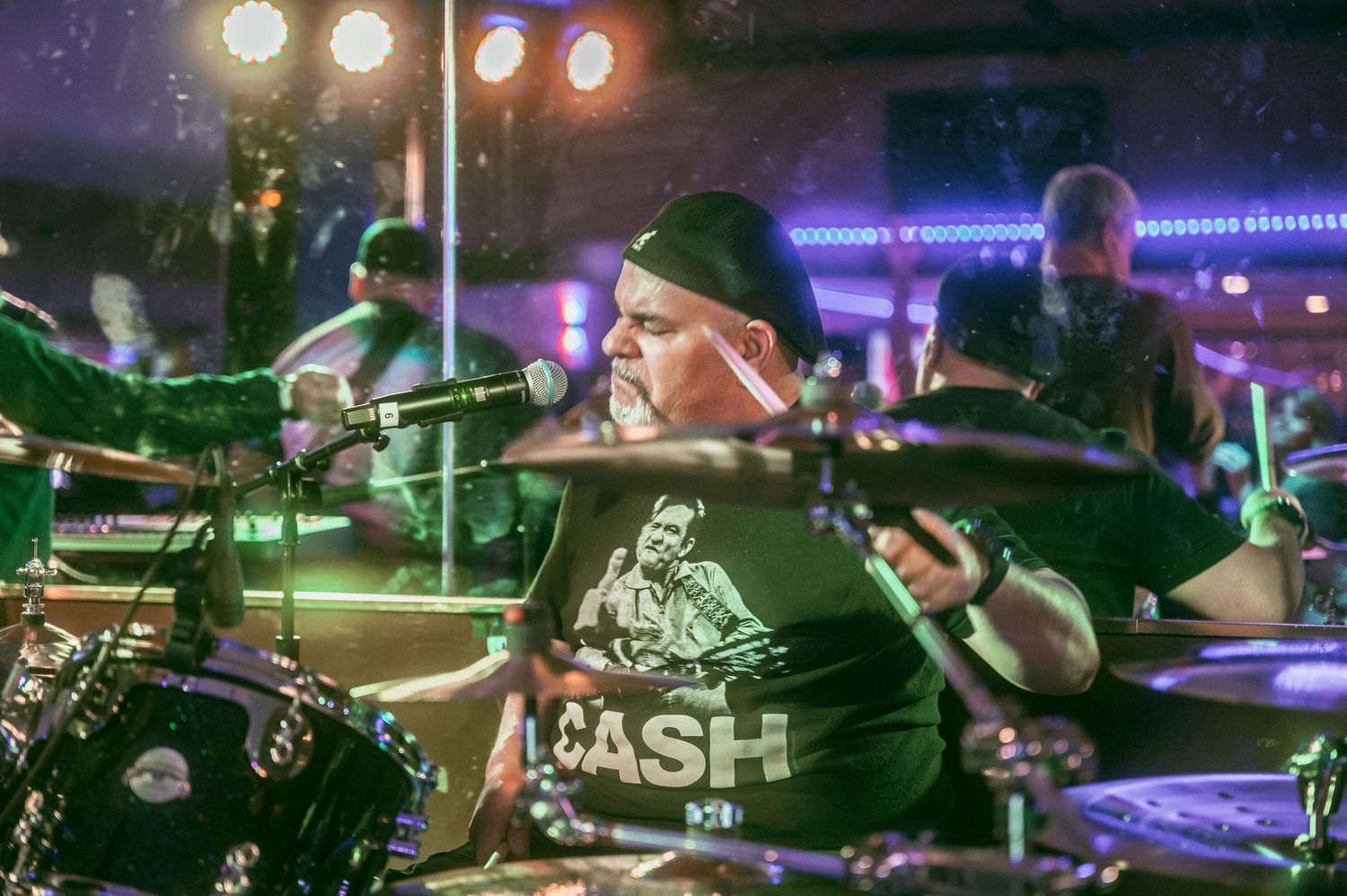 A drummer in a Johnny Cash t-shirt performs on stage under colorful lights at a live music venue.