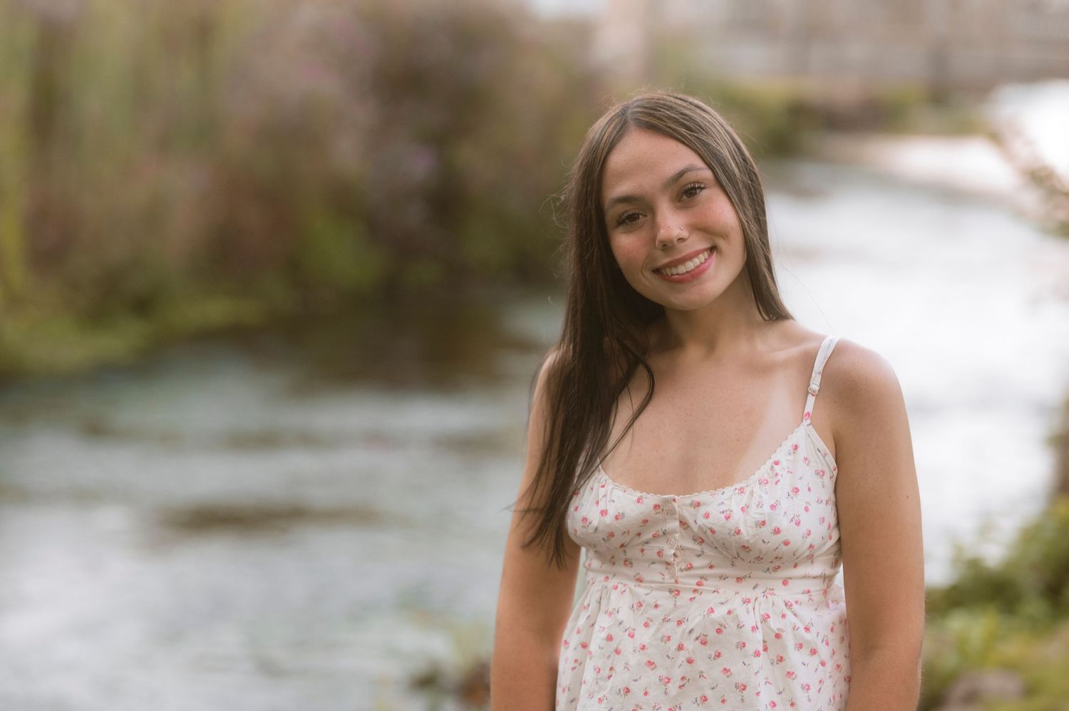 Young woman in floral sundress smiling outdoors near a river with lush greenery in the background.