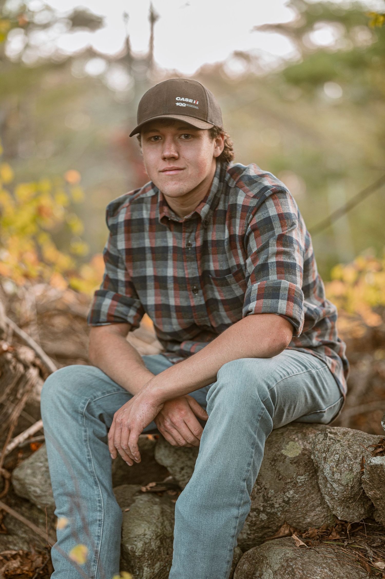 Casual outdoor portrait showing someone in a plaid flannel shirt and baseball cap sitting on rocks in nature.