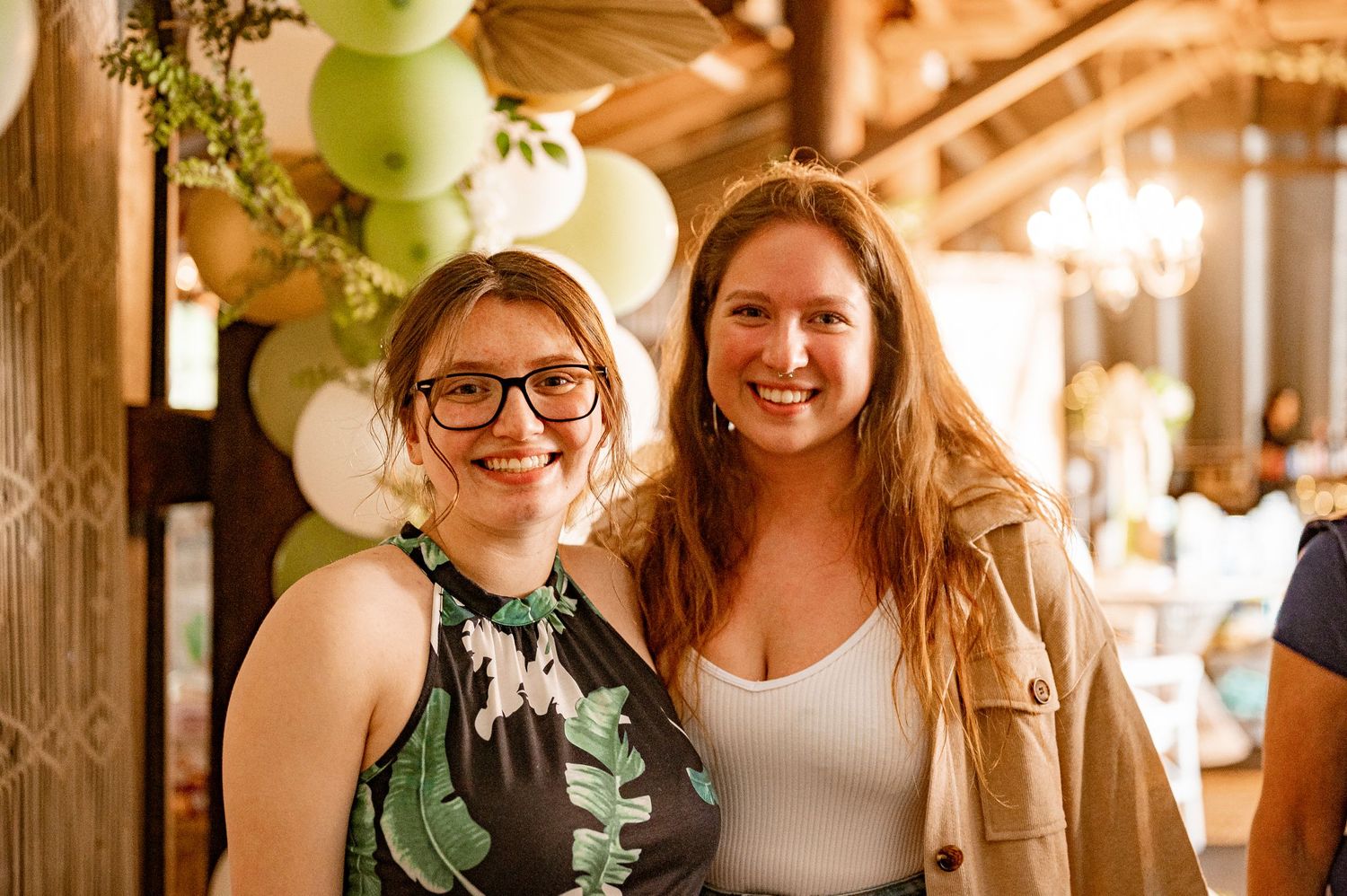 Two friends smile warmly at a party decorated with green balloons and elegant lighting.