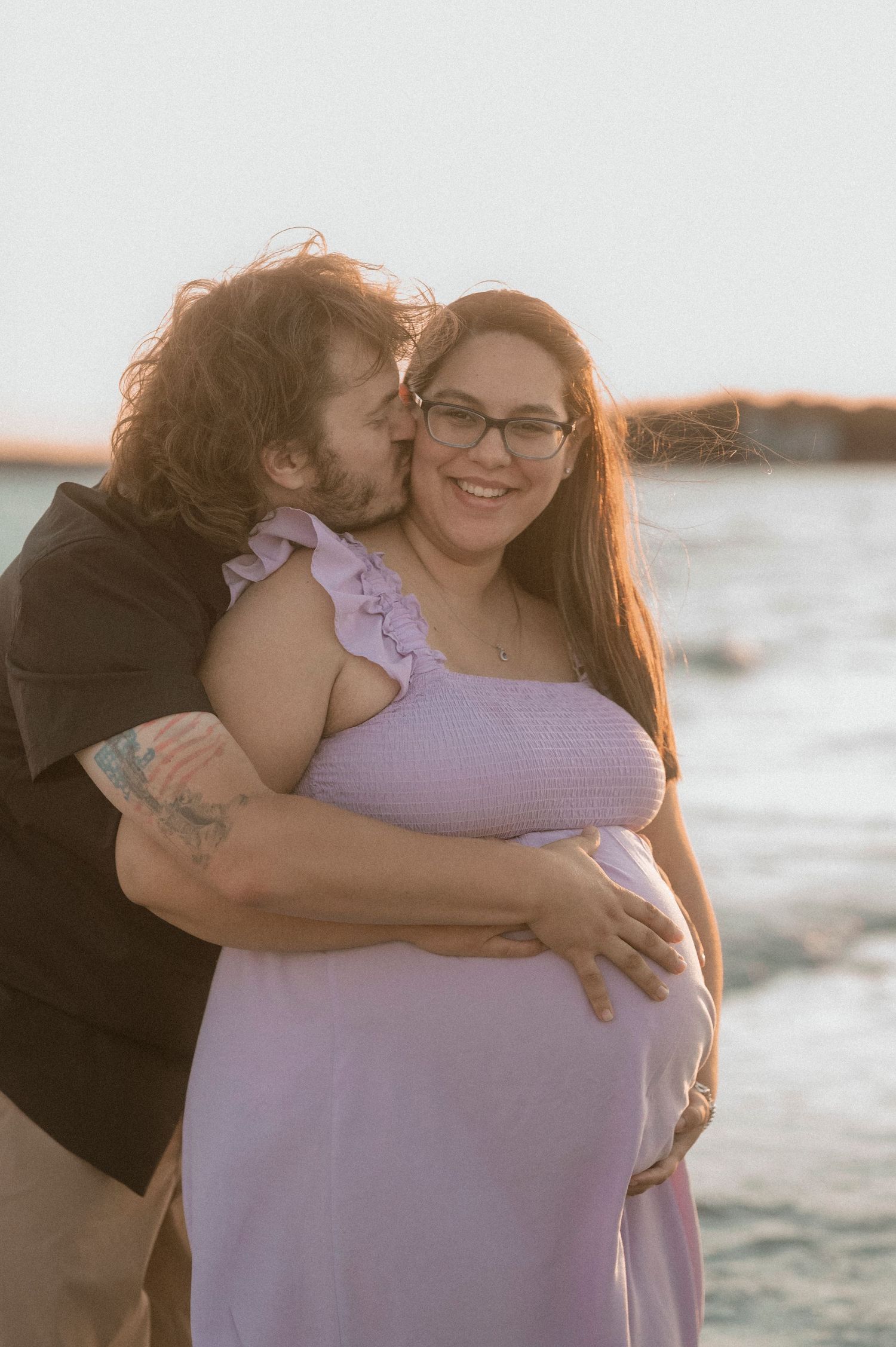 Couple sharing a tender moment at the beach, pregnant woman in lavender dress smiling as partner embraces her at sunset.
