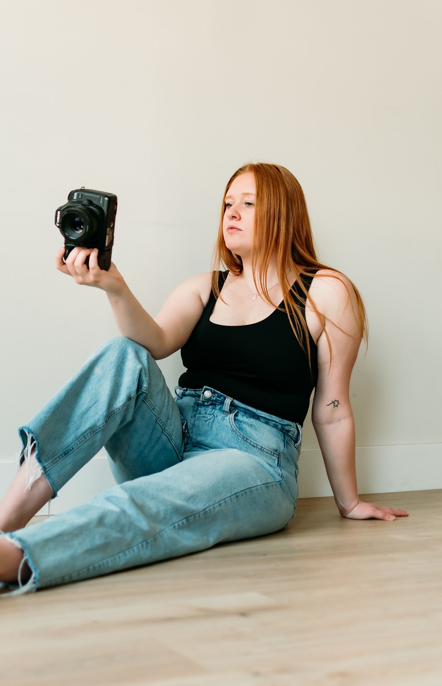 Photographer in black tank top and light blue jeans sits on floor holding vintage instant camera against white wall.