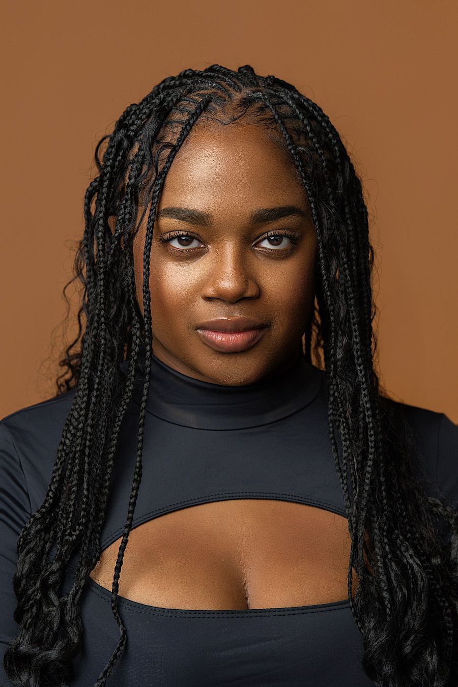 Portrait against brown background wearing black cutout top with long braided hairstyle and natural makeup look.
