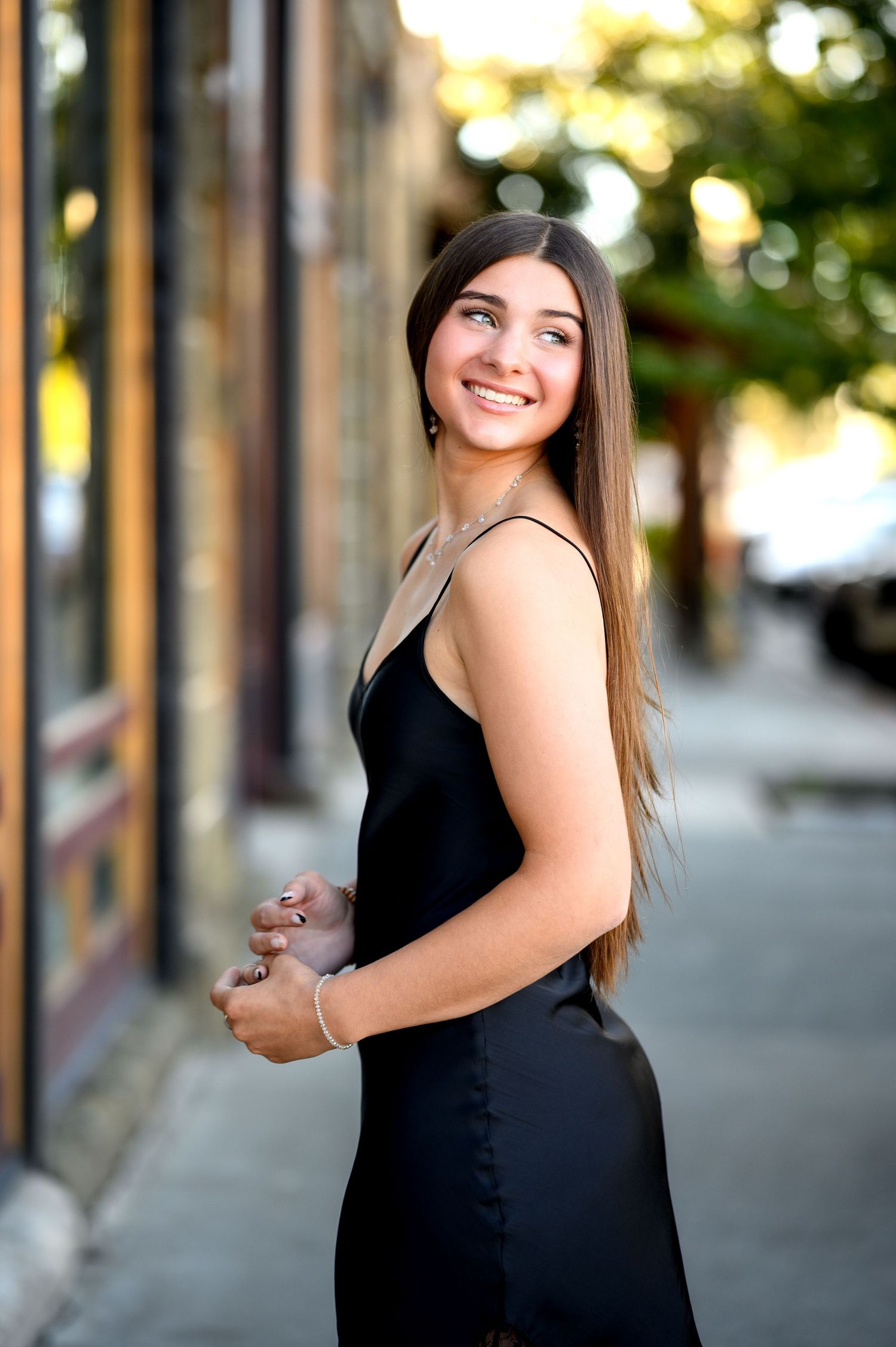 A woman in an elegant black dress smiles warmly while standing on a city sidewalk in soft evening light.