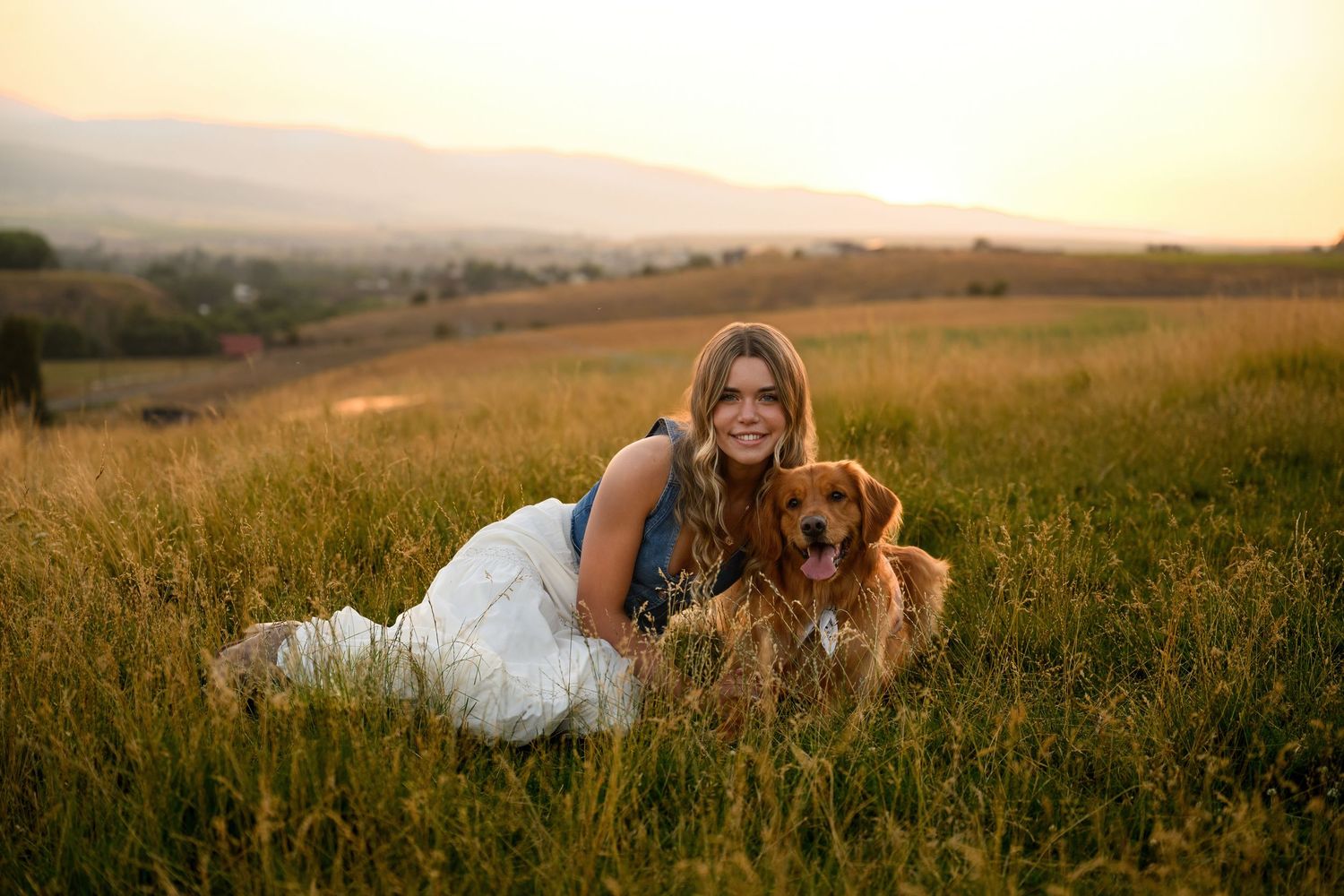 A couple lies together in a golden meadow during sunset while their dog sits nearby watching the horizon.