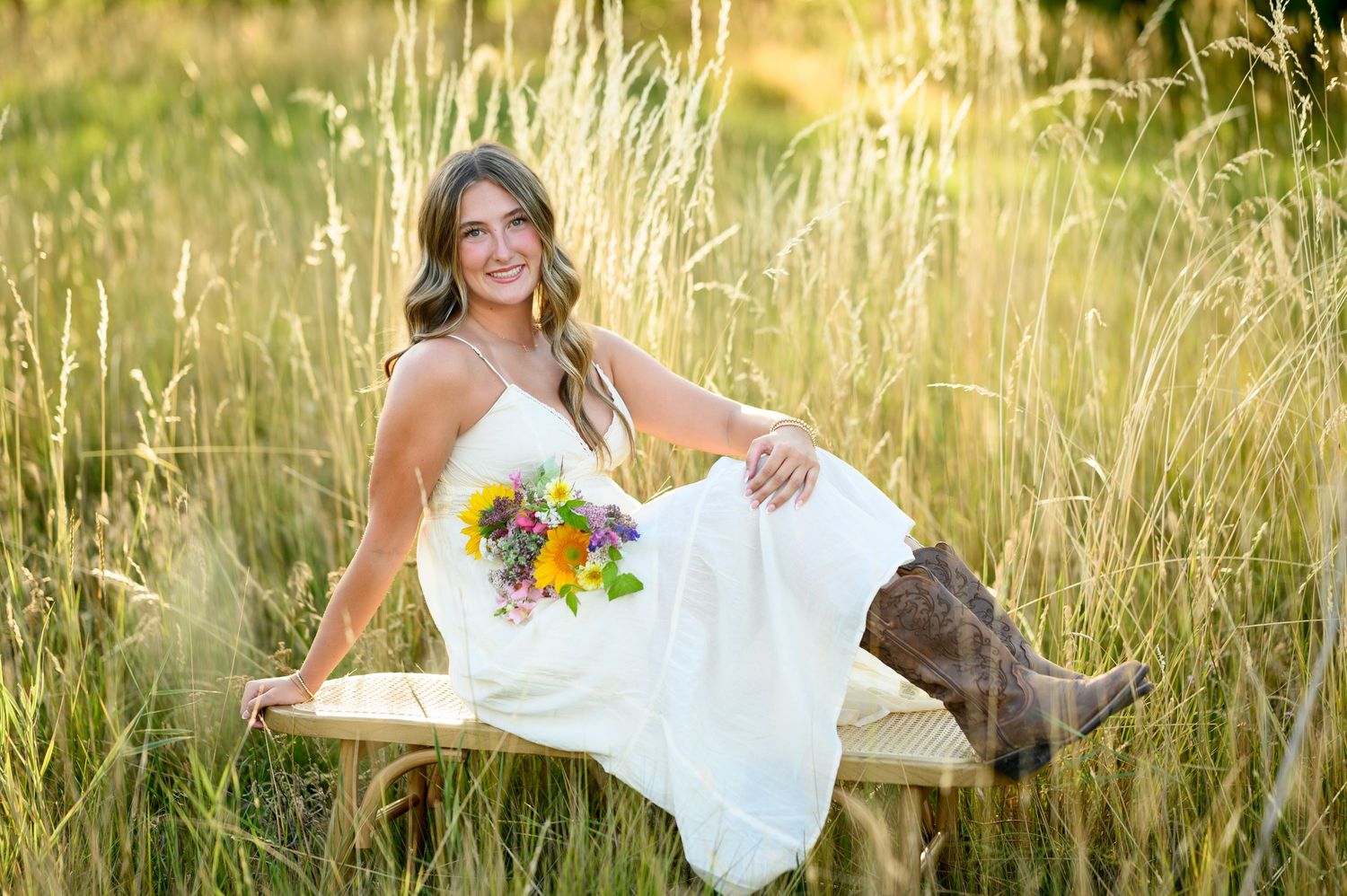 A bride in a white dress and cowboy boots sits on a wooden bench holding sunflowers in a golden wheat field at sunset.