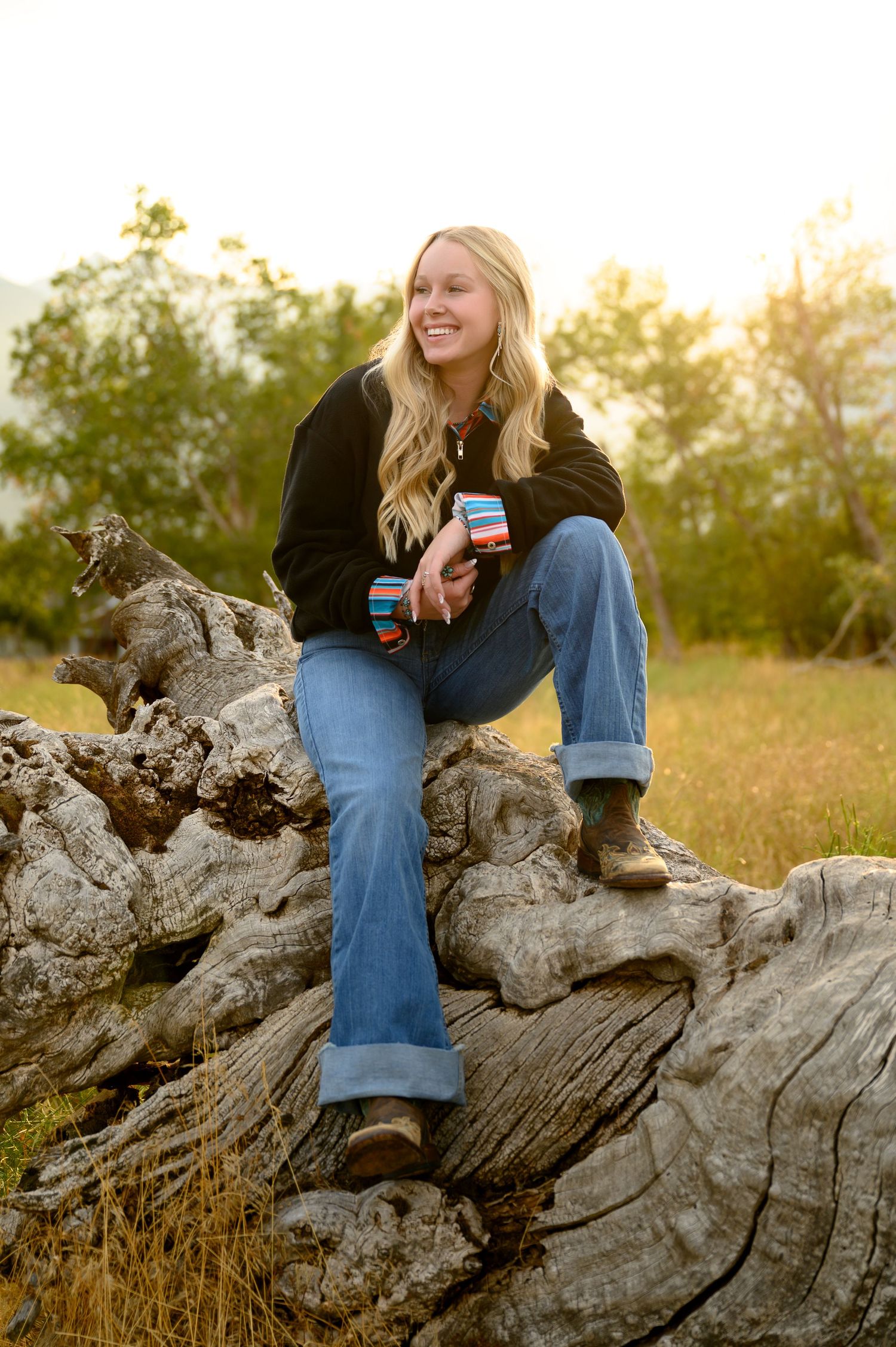 Person in jeans sits on weathered driftwood log at sunset in grassy field holding beverage can.