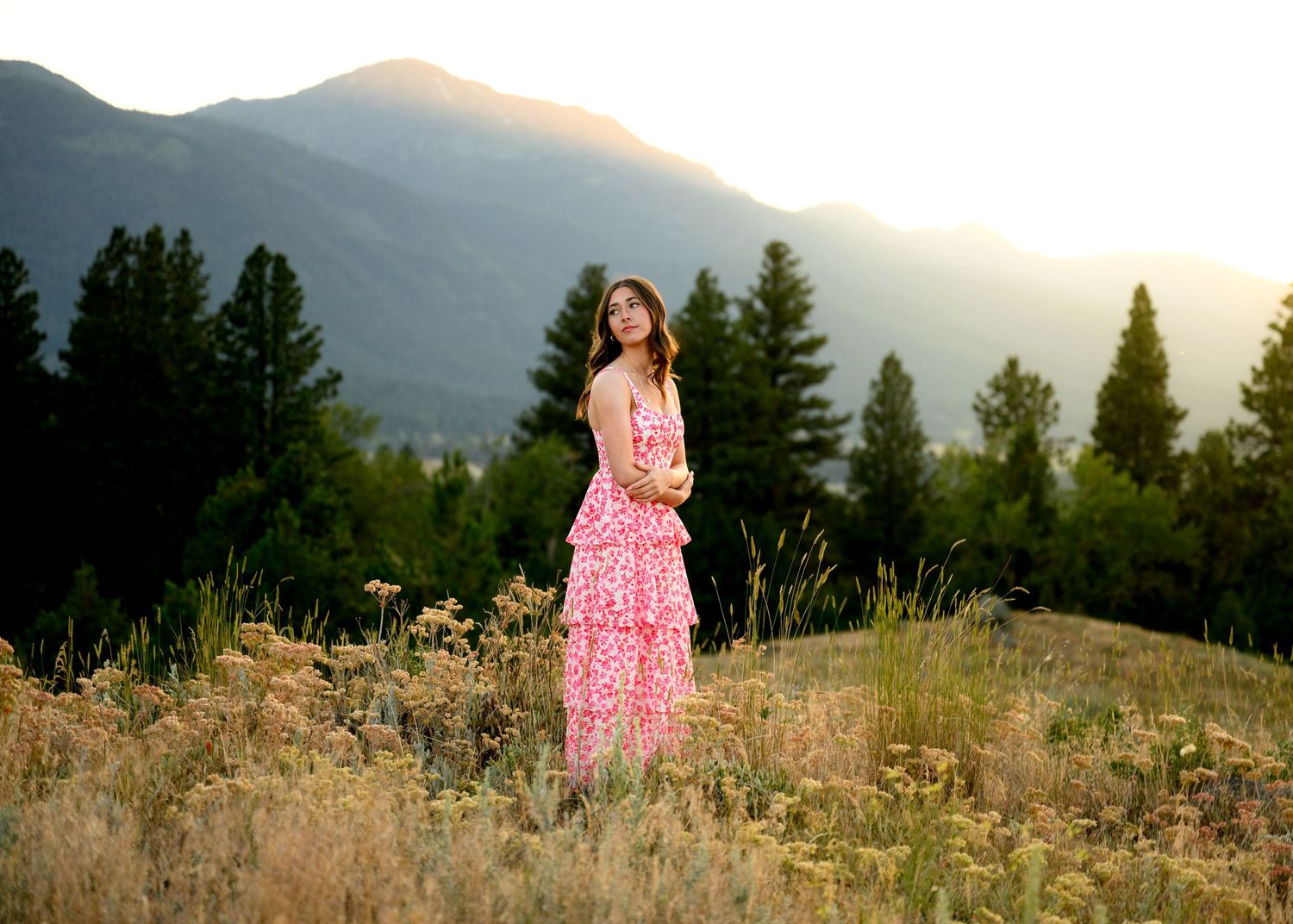 A pink floral dress stands out against a mountain backdrop at golden hour with evergreen trees and wildflowers in view.