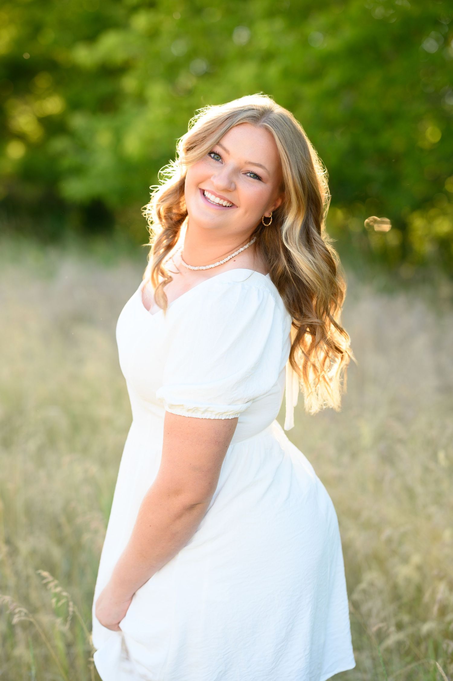 Portrait of someone in a white dress posing outdoors in a sunny field with bokeh effect in the background.