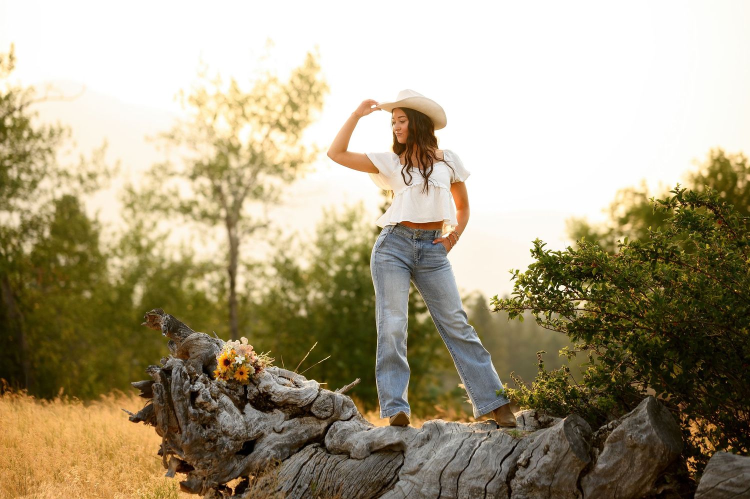 Golden sunset silhouettes trees as someone poses on fallen log in high-waisted jeans and cropped white top.