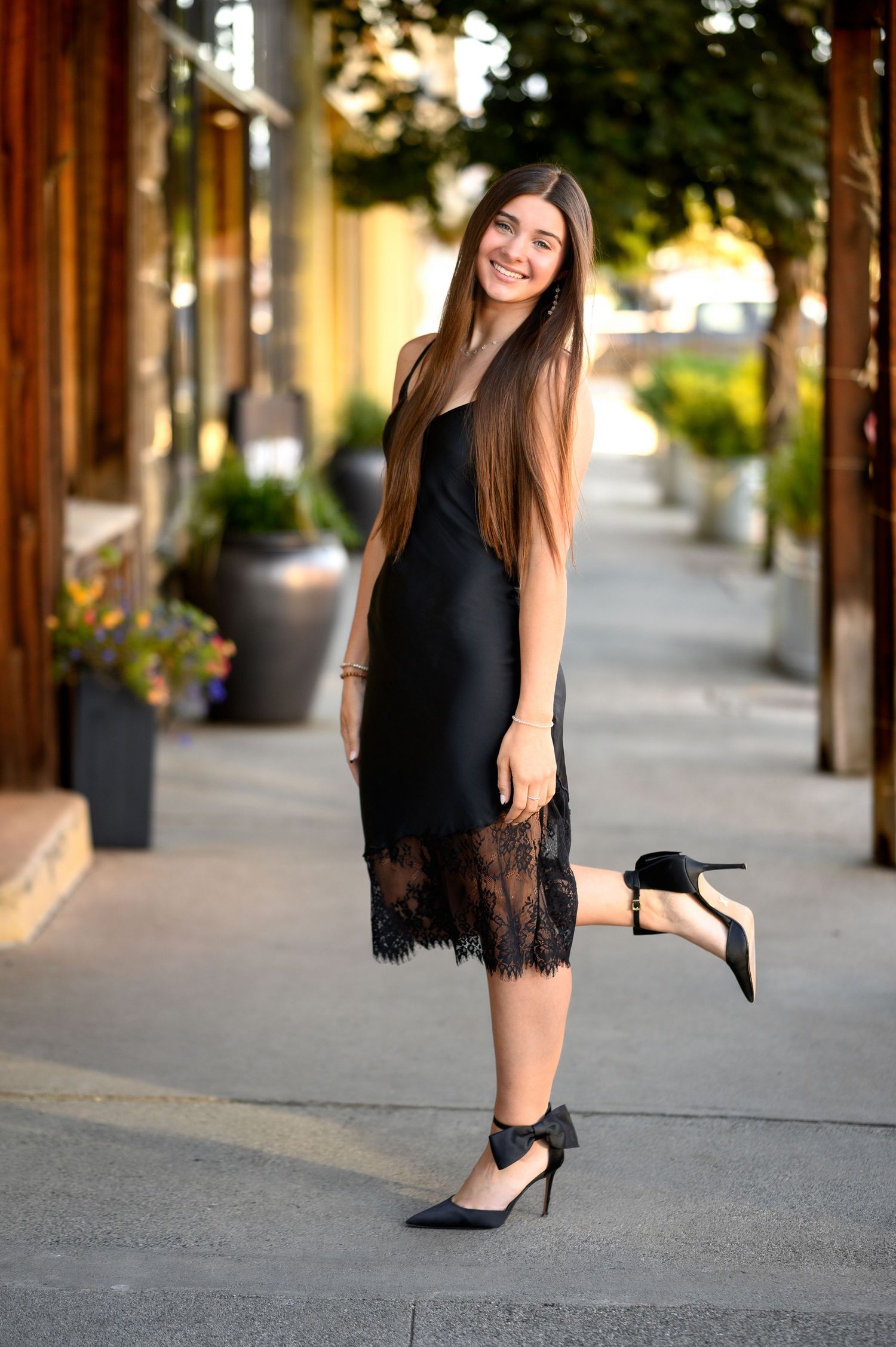 A person in a black lace-trimmed midi dress and heels poses on a sidewalk with potted plants and storefronts in background.