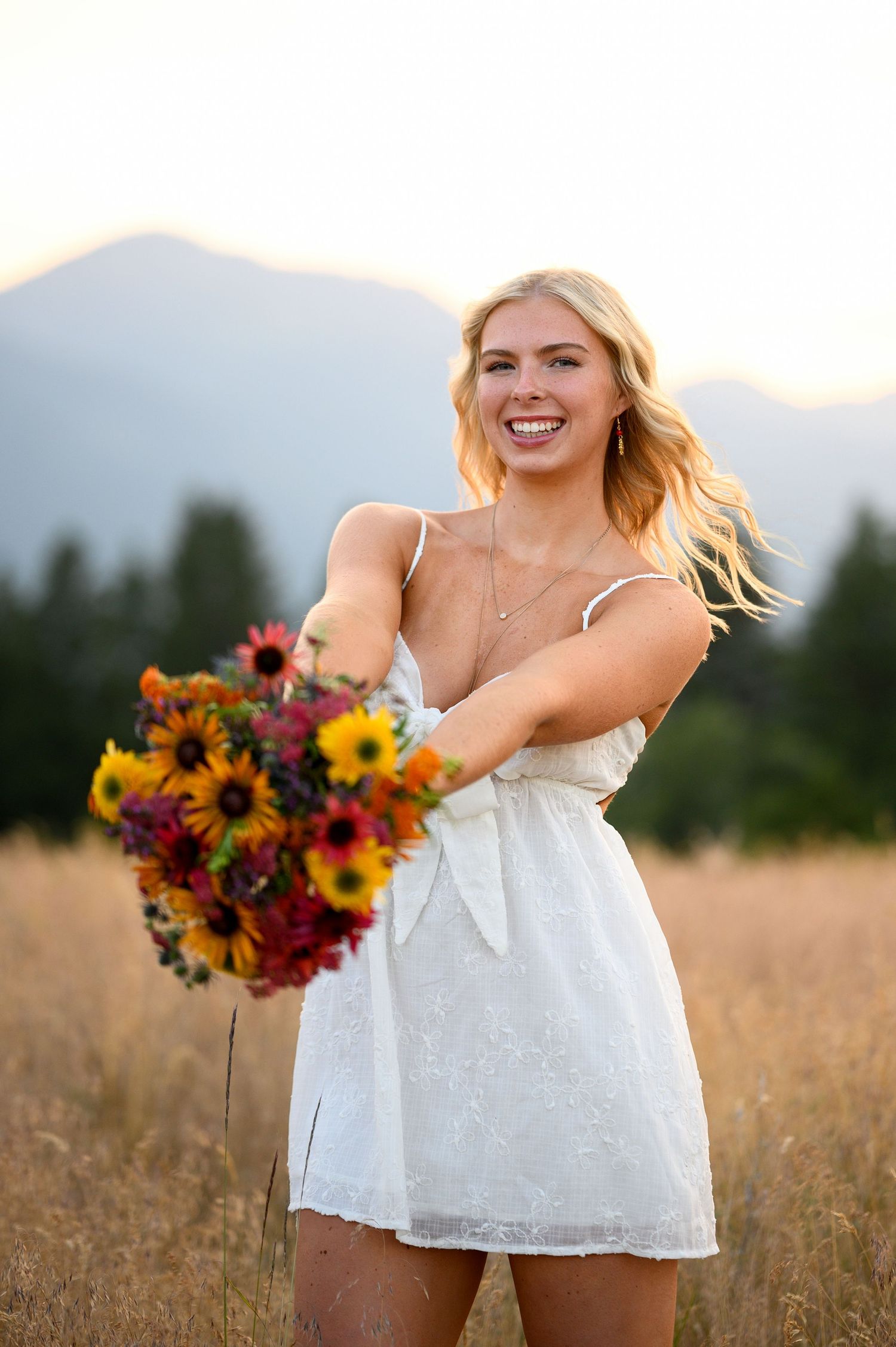 Person in a white sundress holding a vibrant bouquet of sunflowers and wildflowers in a golden field at sunset.