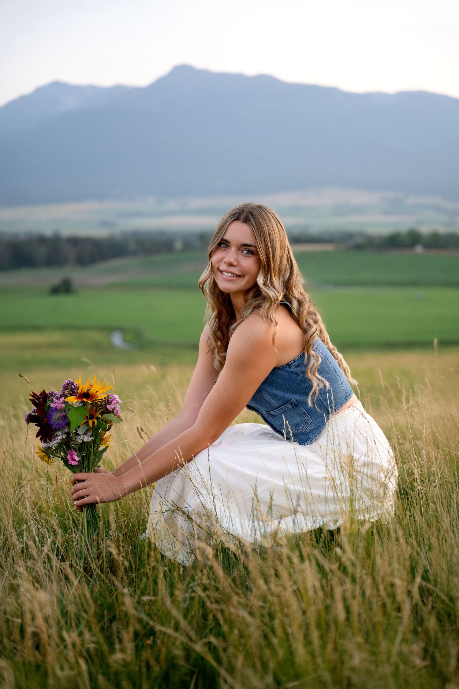 A person in a white dress lies in a grassy meadow holding a colorful bouquet with mountains in the background.
