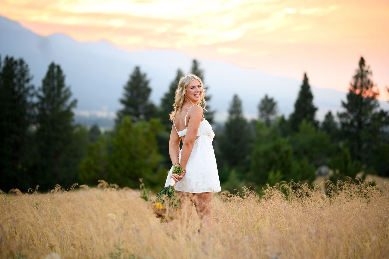 A figure in a white sundress stands in a golden wheat field at sunset with evergreen trees in the background.